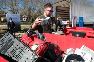 Mężczyzna o jasnej skórze w ciemnych okularach słonecznych wkłada zużyty sprzęt elektroniczny do dużego, czerwonego pojemnika. Ma krótkie, lekko potargane włosy, brodę i założoną ciemnozieloną koszulę. W dłoni trzyma niewielkie, szare urządzenie elektryczne z kablem. Czerwony pojemnik jest wypełniony różnymi elektrośmieciami: widać czarną obudowę prawdopodobnie od monitora lub innego urządzenia, czerwony odkurzacz, słuchawki na pałąku, fragment koła od jakiegoś sprzętu oraz inne plastikowe elementy i kable. W tle stoi otwarta ciężarówka z podniesioną plandeką, do której prowadzi rampa. Przy ciężarówce stoi mężczyzna w czarnej kurtce i spodniach moro. Obok ciężarówki ustawione są dwa pionowe banery reklamowe, jeden biało-czerwony, drugi niebieski z białym napisem „Enea Operator”. Dalej, po lewej stronie, znajduje się tablica lub ścianka informacyjna w odcieniach zieleni z logotypem i tekstem, obok której stoi kobieta w ciemnych ubraniach i okularach przeciwsłonecznych. W tle rosną wysokie, bezlistne drzewa oraz widać fragmenty domów i rabaty z kolorowymi kwiatami. Scena odbywa się na zewnątrz, w słoneczny dzień.