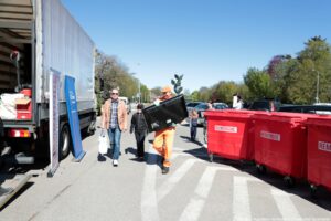 Scena z ulicy w słoneczny dzień. Na pierwszym planie po prawej stronie stoją trzy duże, czerwone kontenery na kółkach z białym napisem „REMONDIS”. Na środku kadru mężczyzna w pomarańczowym stroju roboczym i czapce niesie w ramionach duży płaski telewizor lub monitor, trzymając go ukośnie. Po lewej stronie, bliżej krawędzi, stoi duża ciężarówka z odsuniętą burtą i jasną plandeką; obok ciężarówki ustawione są dwa stojące banery, na jednym widać logo „Enea Operator”. Przed ciężarówką idzie mężczyzna w jasnobrązowej kurtce, dżinsach i okularach przeciwsłonecznych, trzyma biały worek w dłoni; obok niego idzie niższa osoba w ciemnym ubraniu i czapce z daszkiem. W tle po prawej stronie widać zaparkowane samochody i kilka osób, w tym dziecko w kolorowej bluzce, a za nimi drzewa z zielonymi i częściowo czerwonymi liśćmi. Na horyzoncie widać jasne niebo bez chmur i dalszy ciąg ulicy.