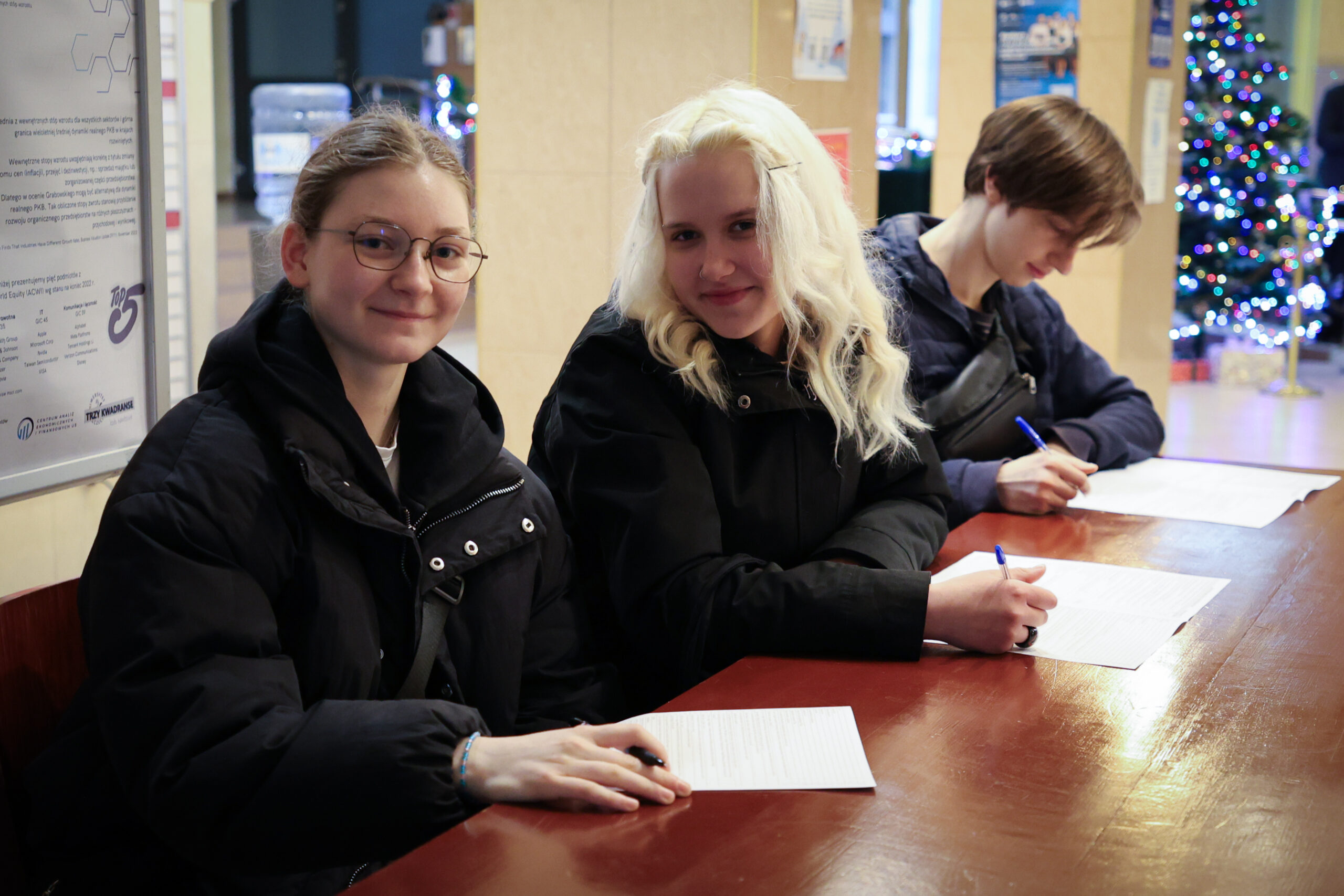 Trzy osoby siedzące przy długim, drewnianym stole i wypełniające kartki papieru długopisami. Po lewej stronie jest młoda kobieta w okularach, z jasną cerą i jasnobrązowymi włosami związanymi do tyłu; ma na sobie czarną kurtkę i lekko się uśmiecha. W środku siedzi młoda kobieta o bardzo jasnych, blond włosach do ramion i jasnej cerze, również w czarnej kurtce; trzyma długopis nad kartką i ma delikatny uśmiech. Po prawej stronie siedzi młoda osoba o jasnej cerze i brązowych włosach opadających na czoło, w ciemnej kurtce; pochyla głowę nad dokumentem i pisze. Na stole leżą trzy zestawy kartek z gęstym tekstem. W tle widać wnętrze budynku, tablicę/informator po lewej oraz choinkę z niebieskimi i białymi światełkami po prawej.