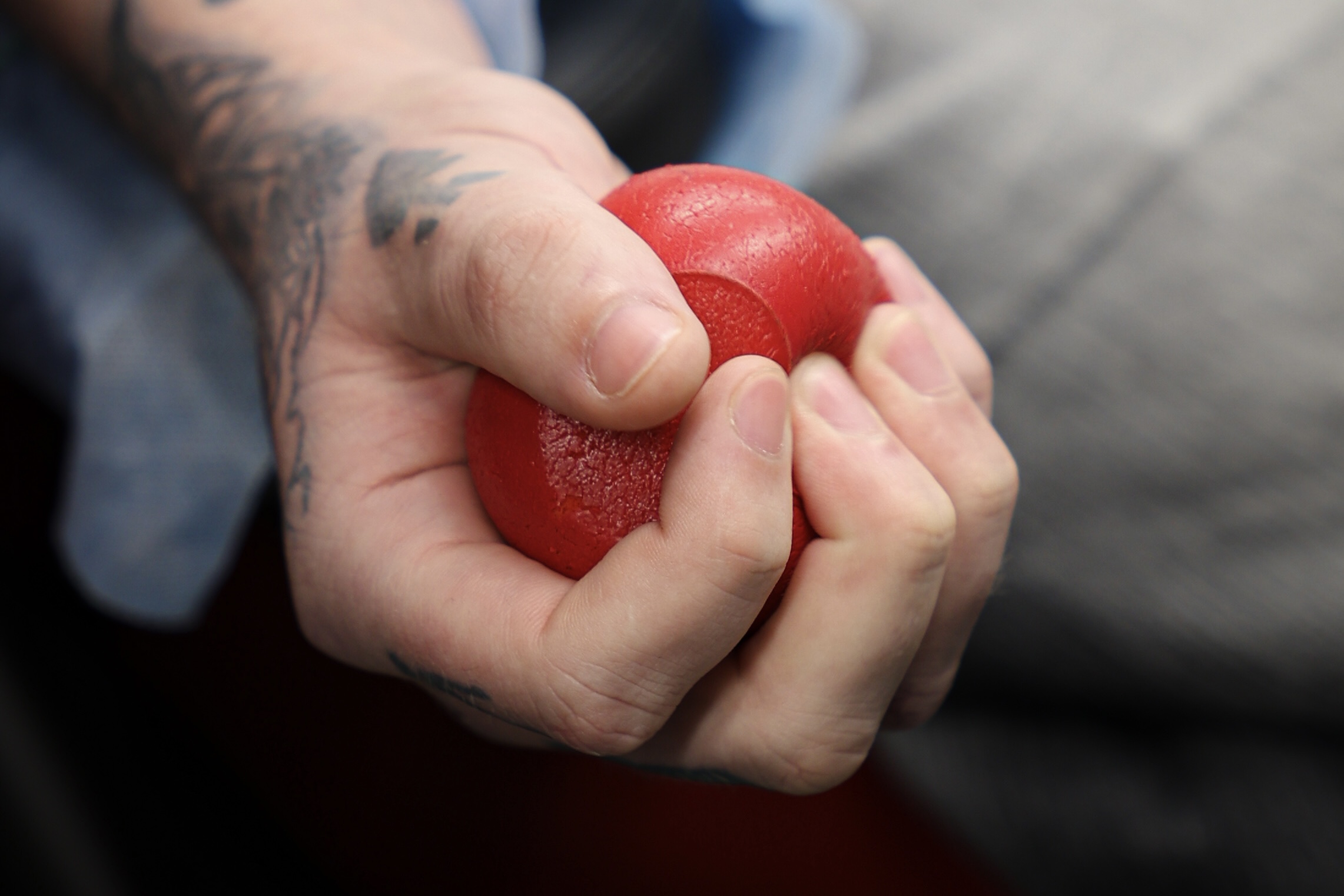 Dłoń o jasnej karnacji ściskająca w środku czerwoną piłeczkę antystresową. Piłeczka jest okrągła, matowa i ma wyraźne wgniecenia od palców. Na skórze przedramienia i przy nadgarstku widać ciemny tatuaż z nieostrymi, roślinnymi lub abstrakcyjnymi kształtami. Tło jest rozmyte, w odcieniach szarości i ciemnego granatu, bez wyraźnych obiektów.