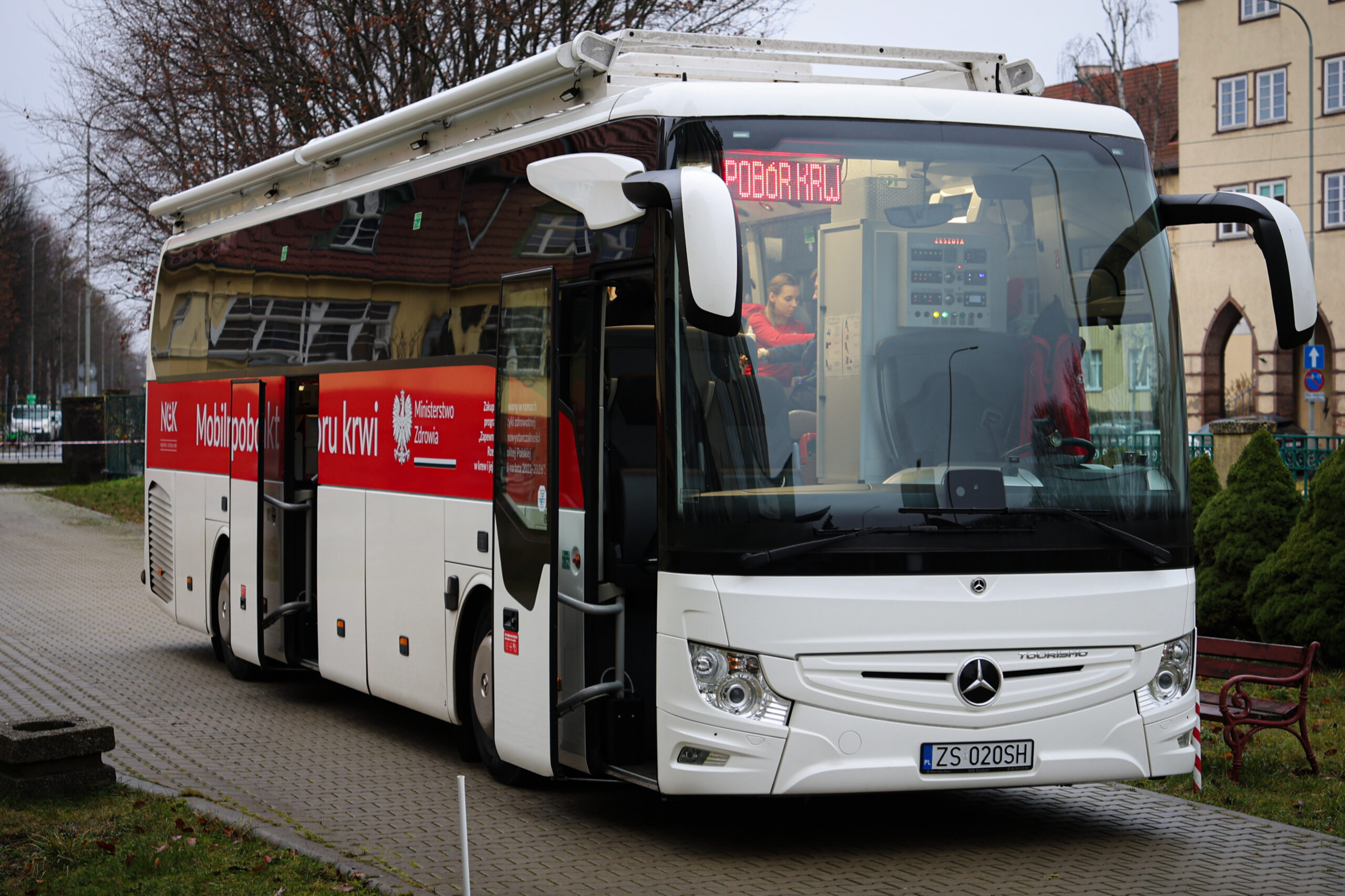 Duży biały autobus typu autokar, zaparkowany na chodniku przy ulicy. Ma czerwony pas na boku z napisami związanymi z krwiodawstwem; na wyświetlaczu z przodu świeci się napis „POBÓR KRWI”. Autobus ma otwarte drzwi po prawej stronie (widać stopnie wejściowe). Z przodu jest logo Mercedesa i tablica rejestracyjna „ZS 020SH”. Na boku w czerwonym pasie da się odczytać m.in. „Mobilnypobor.krwi” oraz „Ministerstwo Zdrowia”; widoczne jest też duże słowo „krwi”. Przez przednią szybę widać wnętrze z panelami urządzeń oraz osobę w czerwonej odzieży pracującą w środku. W tle są budynki, drzewa bez liści i elementy małej architektury (m.in. ławka i krzewy).