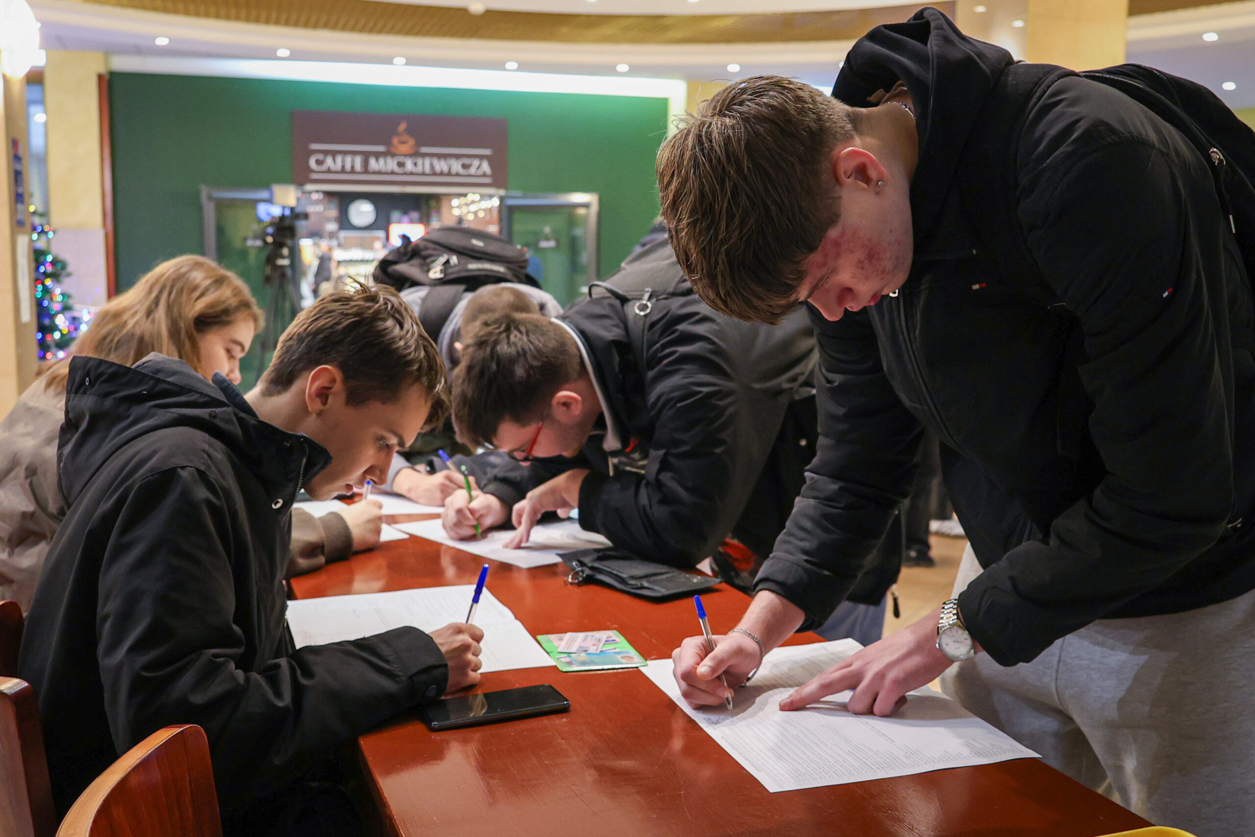 Grupa młodych osób pochylonych nad długim drewnianym stołem, w trakcie wypełniania i podpisywania dokumentów na kartkach papieru. Większość ma na sobie ciemne kurtki i plecaki. Na stole leżą białe formularze, długopisy, czarny smartfon oraz mała zielona ulotka/kartka. Najbliżej po prawej stronie stoi wysoki chłopak w czarnej kurtce i szarych spodniach, pisze długopisem i drugą dłonią przytrzymuje papier; na nadgarstku ma zegarek. Po lewej stronie siedzi chłopak w czarnej kurtce, także pisze na formularzu. W tle widać wnętrze galerii/holu i szyld „CAFFE MICKIEWICZ”.