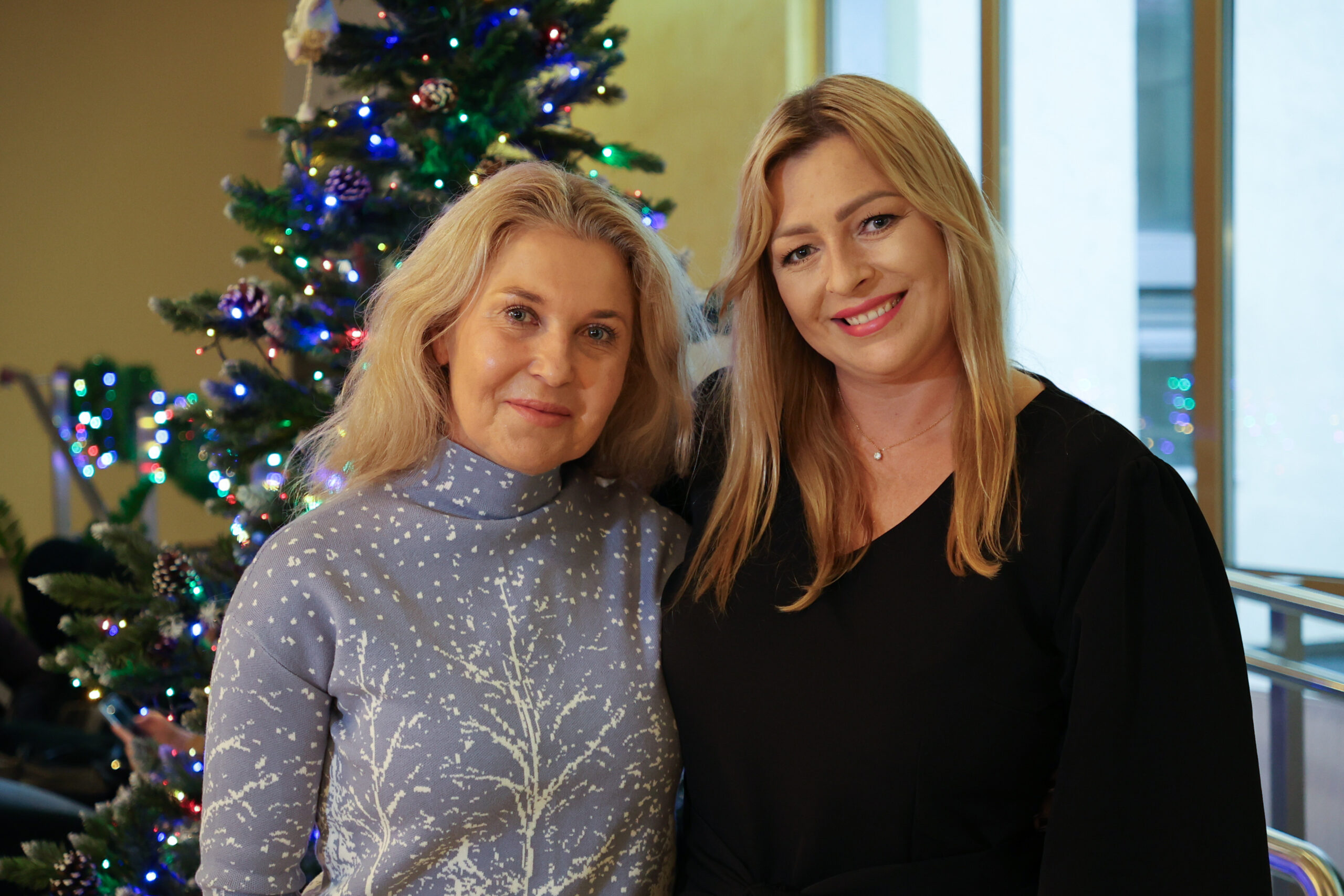 Dwie kobiety stojące blisko siebie na tle udekorowanej choinki z kolorowymi lampkami. Obie mają jasną karnację i blond włosy, uśmiechają się do aparatu. Kobieta po lewej ma włosy do ramion, lekko falowane, rozpuszczone. Ma na sobie jasnofioletowy sweter z wysokim kołnierzem i białym wzorem przypominającym gałęzie lub drzewa. Kobieta po prawej ma dłuższe, proste blond włosy z przedziałkiem na bok. Ma na sobie czarną bluzkę z dekoltem w kształcie litery V i delikatny naszyjnik. W tle widać choinkę z ozdobami i wielokolorowymi światełkami oraz jasne okna sugerujące wnętrze budynku.