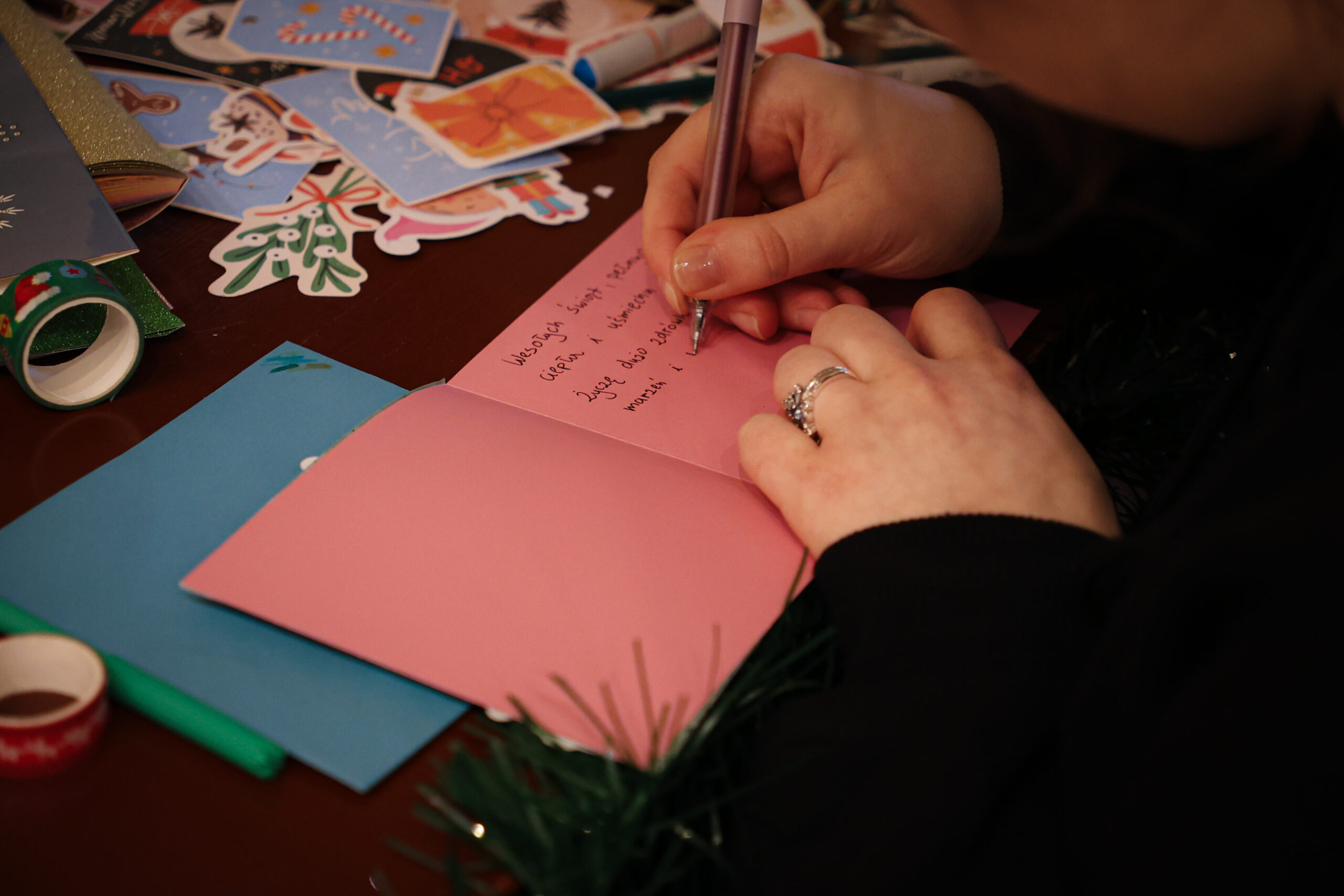 Dłonie piszące długopisem na różowej kartce okolicznościowej rozłożonej na stole. Osoba zapisuje krótką wiadomość w języku polskim; pismo jest częściowo czytelne, ale nie w pełni ostre na całej kartce. Na ręce widać srebrny pierścionek z ozdobnym kamieniem, a skóra dłoni jest jasna. Na stole leżą materiały do robienia kartek i dekoracji: kolorowe wycięte naklejki/papierowe ozdoby (m.in. motywy świąteczne), rolka zielonej taśmy dekoracyjnej, niebieska koperta lub kartka, zielony marker oraz inne przybory. W dolnej części kadru widać fragment zielonej, iglastej dekoracji przypominającej gałązkę choinki.