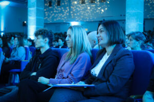 Trzy osoby siedzące w pierwszym rzędzie na widowni sali konferencyjnej o niebieskim oświetleniu. W tle widać wiele innych osób na miejscach, kolumny oraz świetlne punkty na ścianie i suficie, jak z projektora lub dekoracyjnego oświetlenia. Po lewej siedzi młody mężczyzna o jasnej skórze, kręconych ciemnych włosach i okularach, w ciemnej bluzie; jest zwrócony bokiem i patrzy w stronę sceny. Na środku siedzi kobieta o jasnej skórze i długich prostych blond włosach, w fioletowej bluzie; ma na szyi smycz z identyfikatorem i także jest skierowana w stronę sceny. Po prawej siedzi kobieta o jasnej skórze, z ciemnymi włosami do ramion, w ciemnym żakiecie i białej bluzce; trzyma na kolanach teczkę lub dokumenty i lekko się uśmiecha, również patrząc w stronę sceny.