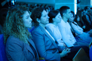 Rząd osób siedzących obok siebie na widowni podczas wydarzenia w sali. Dominują chłodne, niebieskie barwy oświetlenia. Na pierwszym planie po lewej stronie siedzi kobieta o jasnej skórze i jasnobrązowych, bardzo kręconych włosach do ramion, w marynarce we wzór. Obok niej siedzi kobieta o jasnej skórze, z ciemnymi włosami związanymi z tyłu, w szarej marynarce; ma ręce złożone na kolanach. Dalej widać młodą osobę o jasnej skórze w okularach, w jasnej bluzie. Kolejna osoba to młody mężczyzna o jasnej skórze w białej koszuli, siedzący prosto i patrzący w tę samą stronę co reszta. W tle po prawej stronie widocznych jest więcej uczestników, siedzących w rzędach i skierowanych w stronę sceny poza kadrem.