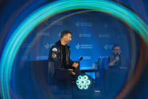 Scena z panelu dyskusyjnego na tle niebieskiej ścianki z powtarzającym się napisem „UNIWERSYTET SZCZECIŃSKI” i logo „US”. Po lewej siedzi mężczyzna o jasnej skórze i krótkich ciemnych włosach, uśmiecha się i trzyma mikrofon; ma na sobie czarną kurtkę (z naszywkami na rękawie) i czarne spodnie. Po prawej siedzi kobieta o jasnej skórze w okularach, trzyma mikrofon blisko ust. Między nimi stoi niski stolik z kilkoma szklankami i butelkami. Na dole kadru jest widoczny reflektor sceniczny świecący na biało, a cały obraz ma efekt kolorowej, okrągłej poświaty/obręczy wokół krawędzi.