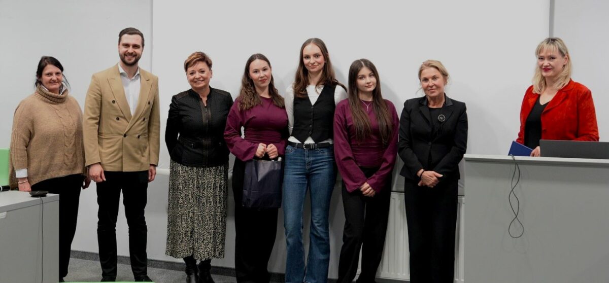 Grupa ośmiu osób stojących w rzędzie na tle białej ściany w sali wykładowej. To 7 kobiet i 1 mężczyzna, wszyscy patrzą w stronę aparatu. Większość ma jasną karnację; jedna kobieta po lewej ma ciemniejsze włosy i również wygląda na jasnokarną. Ubrania są eleganckie lub półformalne: beże, czerń oraz odcienie bordo i fioletu. Mężczyzna (drugi od lewej) ma beżową marynarkę. Dwie młodsze kobiety w środku mają bordowe bluzki; jedna z nich trzyma ciemną torbę. Kobieta skrajnie po prawej ma czerwony żakiet i stoi przy mównicy z laptopem; widać też zwisające kable.