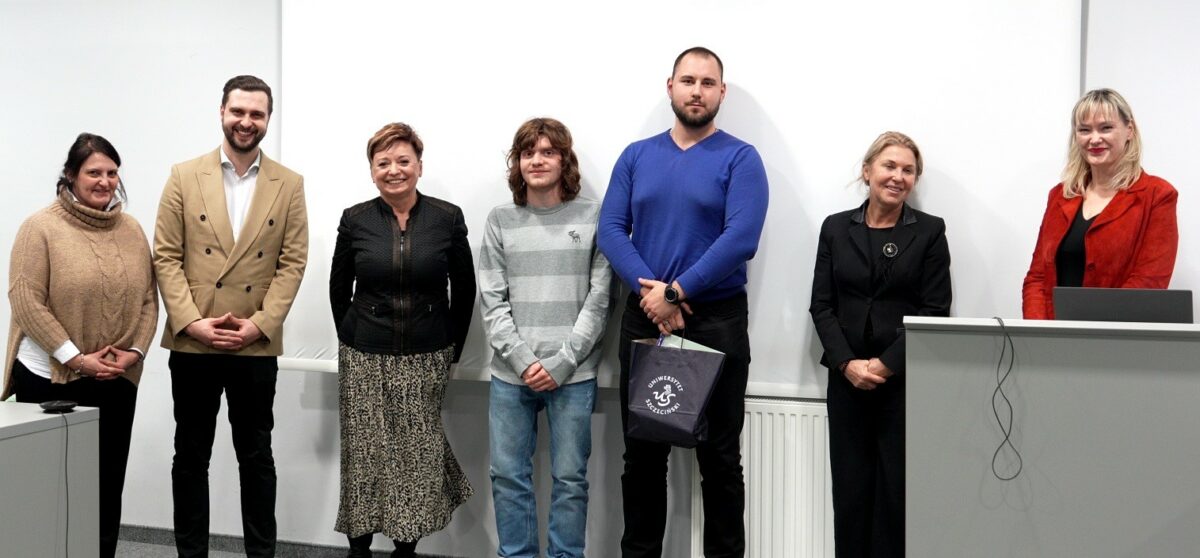 Grupa siedmiu osób stojących w jednym rzędzie na tle białej ściany w sali. To zdjęcie wygląda na formalne, zrobione podczas spotkania lub wydarzenia. Wszystkie osoby są dorosłe i uśmiechają się lub mają neutralny wyraz twarzy. Od lewej: kobieta w beżowym swetrze z golfem. Mężczyzna w jasnobeżowej, dwurzędowej marynarce i czarnych spodniach. Kobieta w czarnej górze i długiej, wzorzystej spódnicy. Młody mężczyzna w szaro-białym, pasiastym swetrze i jasnych dżinsach. Wysoki mężczyzna w niebieskim swetrze trzyma ciemną torbę z białym nadrukiem. Kobieta w czarnym stroju. Kobieta w czerwonej marynarce stoi za szarym podium z przewieszonym kablem.