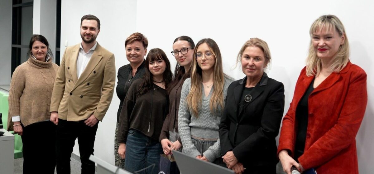 Grupa ośmiu osób stojących w jednym rzędzie na tle białej ściany, pozujących do zdjęcia. Wszyscy patrzą w stronę aparatu i większość się uśmiecha. Od lewej: kobieta w beżowym, grubym swetrze z golfem; mężczyzna z krótką brodą w jasnobeżowej, dwurzędowej marynarce i białej koszuli; kobieta z krótkimi, rudawymi włosami w ciemnym stroju; młoda kobieta z długimi, ciemnymi włosami i grzywką w ciemnej bluzce; kobieta w okularach z ciemnymi włosami związanymi z tyłu, w ciemnym stroju; młoda kobieta w okularach, z długimi włosami, w jasnoszarym swetrze; starsza kobieta w czarnej marynarce z ozdobną broszką; kobieta z jasnymi blond włosami w czerwonej marynarce i czarnym topie.