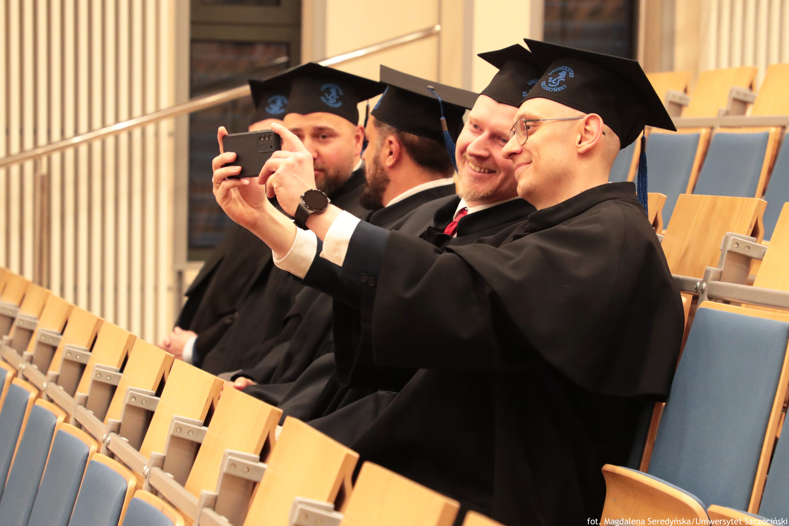 Grupa mężczyzn w czarnych togach i czarnych biretach siedząca w rzędzie na sali wykładowej. Najbliżej aparatu siedzi mężczyzna w okularach, z jasną karnacją, który wyciąga ręce do przodu i robi zdjęcie smartfonem; na ręku ma ciemny zegarek. Obok niego siedzi uśmiechnięty mężczyzna o jasnej karnacji w todze, z widocznym czerwonym krawatem. Dalej w rzędzie są kolejne dwie osoby w podobnych strojach akademickich, częściowo zasłonięte. W tle widać rzędy drewnianych siedzeń z niebieskimi oparciami oraz jasne ściany sali. W prawym dolnym rogu jest napis: „fot. Magdalena Seredyńska/Uniwersytet Szczeciński”.