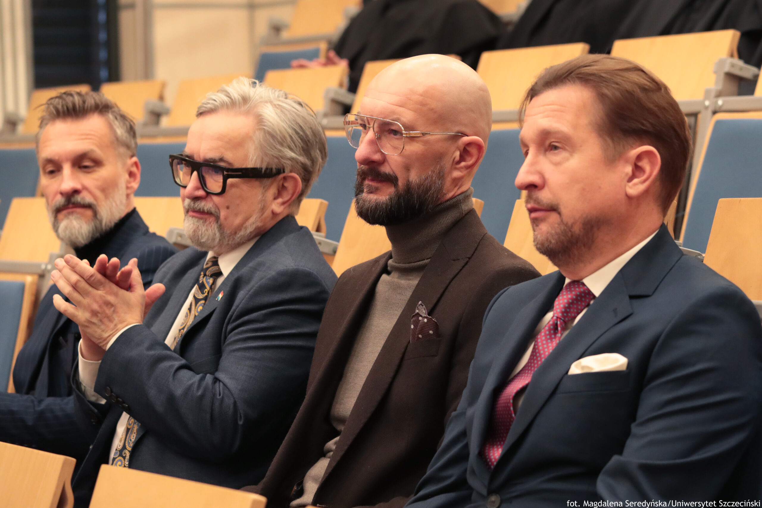 Czterech mężczyzn siedzących w pierwszym rzędzie sali wykładowej. Wszyscy mają jasną karnację i są ubrani formalnie w marynarki. Od lewej: Mężczyzna w średnim wieku z krótkimi, ciemnoszarymi włosami i siwą brodą, w ciemnej marynarce. Starszy mężczyzna z siwymi włosami zaczesanymi do tyłu, w dużych czarnych okularach, z siwą brodą i wąsami; ma niebieską marynarkę i wzorzysty krawat, dłonie ma złożone jak do oklasków. Mężczyzna w średnim wieku, łysy, z ciemną brodą, w okularach; ma ciemną marynarkę i szary golf, w kieszonce poszetka. Mężczyzna w średnim wieku z brązowymi włosami zaczesanymi na bok i lekkim zarostem; ma granatowy garnitur, białą koszulę i czerwony krawat oraz jasną poszetkę. W tle widać rzędy jasnych, drewnianych siedzisk z niebieskimi oparciami, jak w auli. W prawym dolnym rogu jest mały napis: „fot. Magdalena Seredyńska/Uniwersytet Szczeciński”.