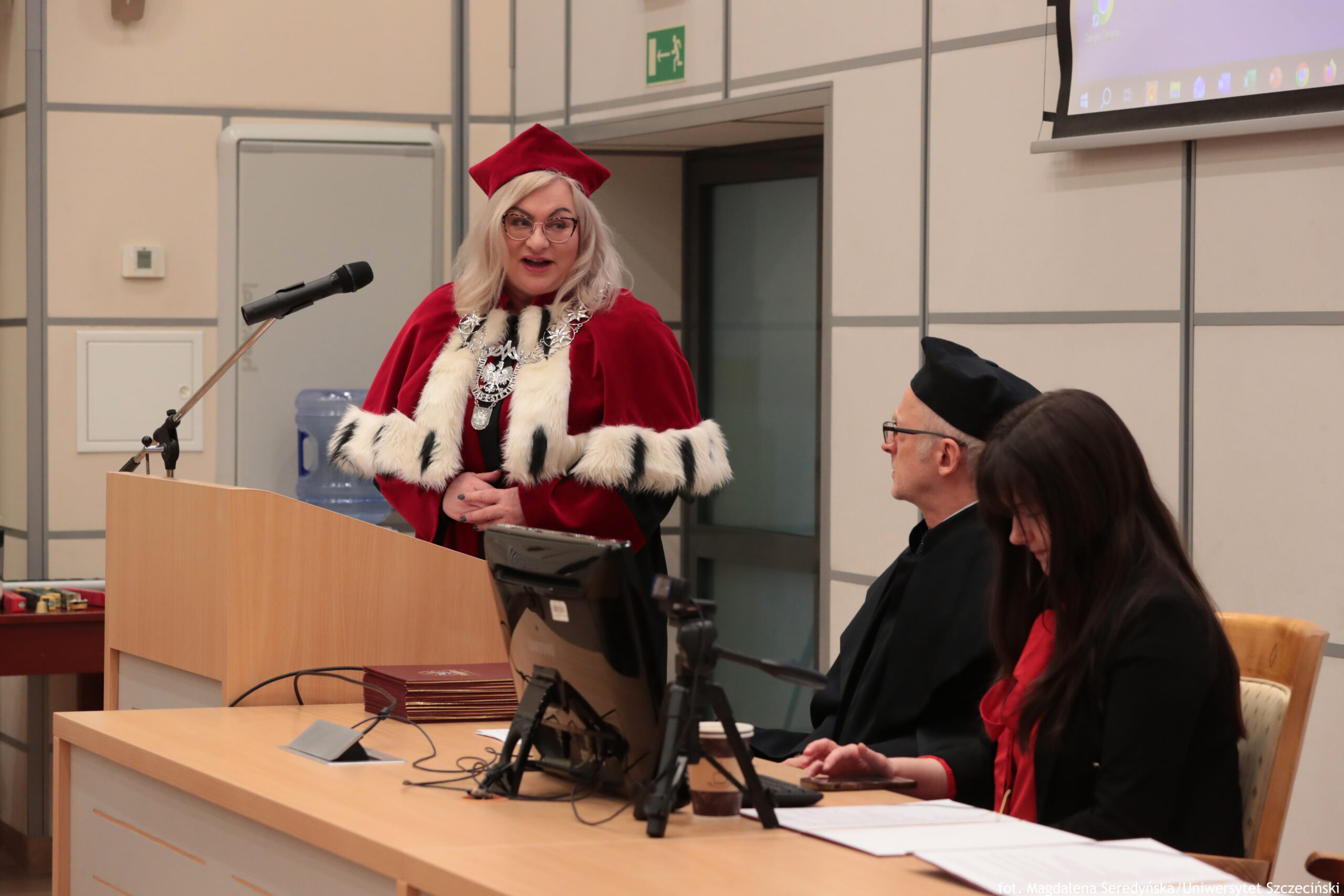 Sala wykładowa podczas uroczystości akademickiej. Na pierwszym planie kobieta o jasnej karnacji i jasnych, blond włosach stoi przy mównicy z mikrofonem, ubrana w czerwony strój ceremonialny z białym futrzanym obszyciem w czarne plamy oraz czerwony biret; na szyi ma ozdobny łańcuch. Po prawej stronie, przy stole, siedzą dwie osoby: starszy mężczyzna w czarnym stroju i czapce akademickiej, w okularach, oraz kobieta z długimi, ciemnymi włosami w czarnym ubraniu z czerwonym akcentem, pochylona nad dokumentami. Na stole stoi monitor komputera, przewody, dokumenty i mały statyw. W tle widać drzwi z zielonym znakiem ewakuacyjnym oraz fragment ekranu projekcyjnego w prawym górnym rogu. W prawym dolnym rogu zdjęcia jest mały podpis, ale tekst jest zbyt drobny, by dało się go pewnie odczytać.