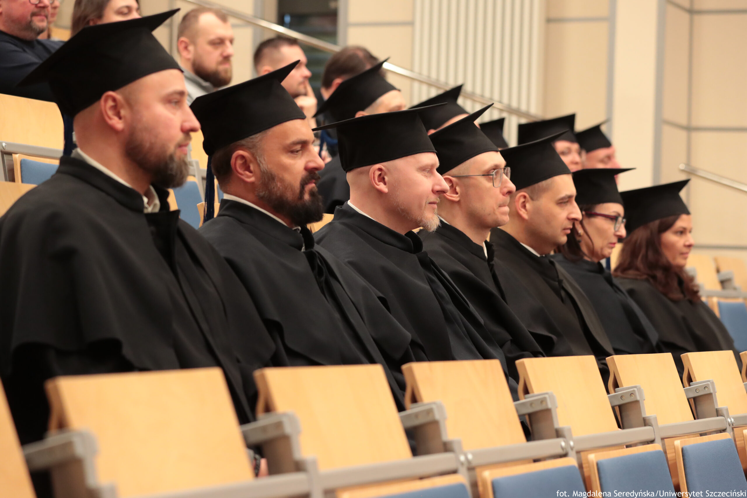 Rząd osób w czarnych togach i biretach siedzących w auli. Widać głównie profil kilku mężczyzn i jednej kobiety, ustawionych obok siebie, wszyscy skierowani w tę samą stronę, jak podczas ceremonii akademickiej. Większość ma jasną karnację; część ma brody lub zarost, jedna osoba ma okulary. Na pierwszym planie są jasne, drewniane oparcia siedzeń, a w tle kolejne rzędy miejsc i kilka osób bez biretów. W prawym dolnym rogu znajduje się mały podpis: „fot. Magdalena Seredyńska/Uniwersytet Szczeciński”.