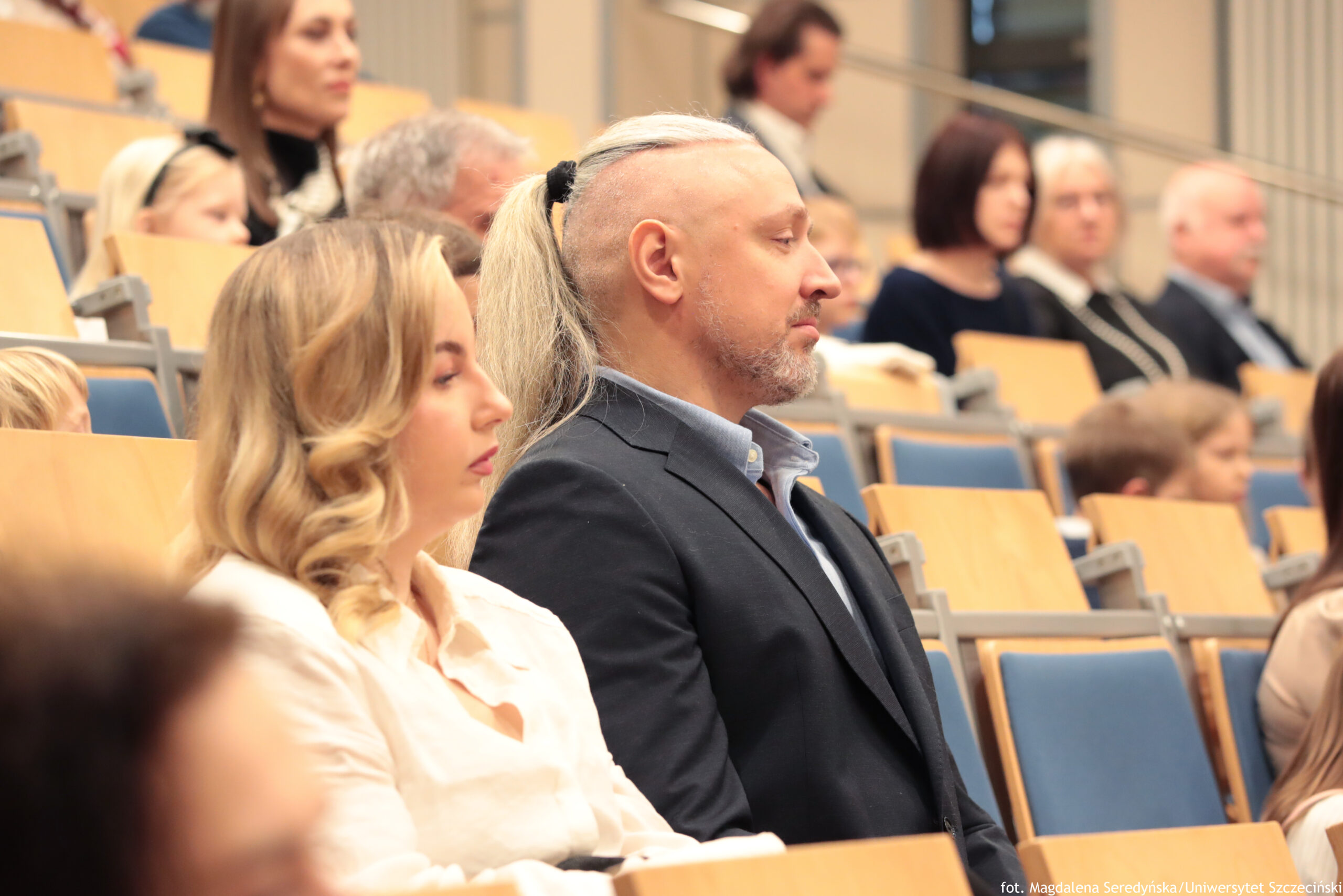 Grupa osób siedzących w sali wykładowej na rzędach jasnych, drewnianych siedzeń z niebieskimi oparciami. W pierwszym planie widać dwie osoby siedzące obok siebie: młoda kobieta o jasnej skórze i blond, falowanych włosach w jasnej koszuli oraz mężczyzna o jasnej skórze w ciemnej marynarce i jasnej koszuli, z wygolonymi bokami głowy i długimi, jasnymi włosami związanymi z tyłu; ma krótki zarost. Oboje mają neutralne, skupione wyrazy twarzy i są zwróceni w tę samą stronę, jak podczas słuchania wystąpienia. W tle siedzi więcej osób w różnych rzędach, większość w stonowanych, formalnych ubraniach; tło jest lekko rozmyte. W prawym dolnym rogu znajduje się mały napis: „fot. Magdalena Seredyńska / Uniwersytet Szczeciński”.