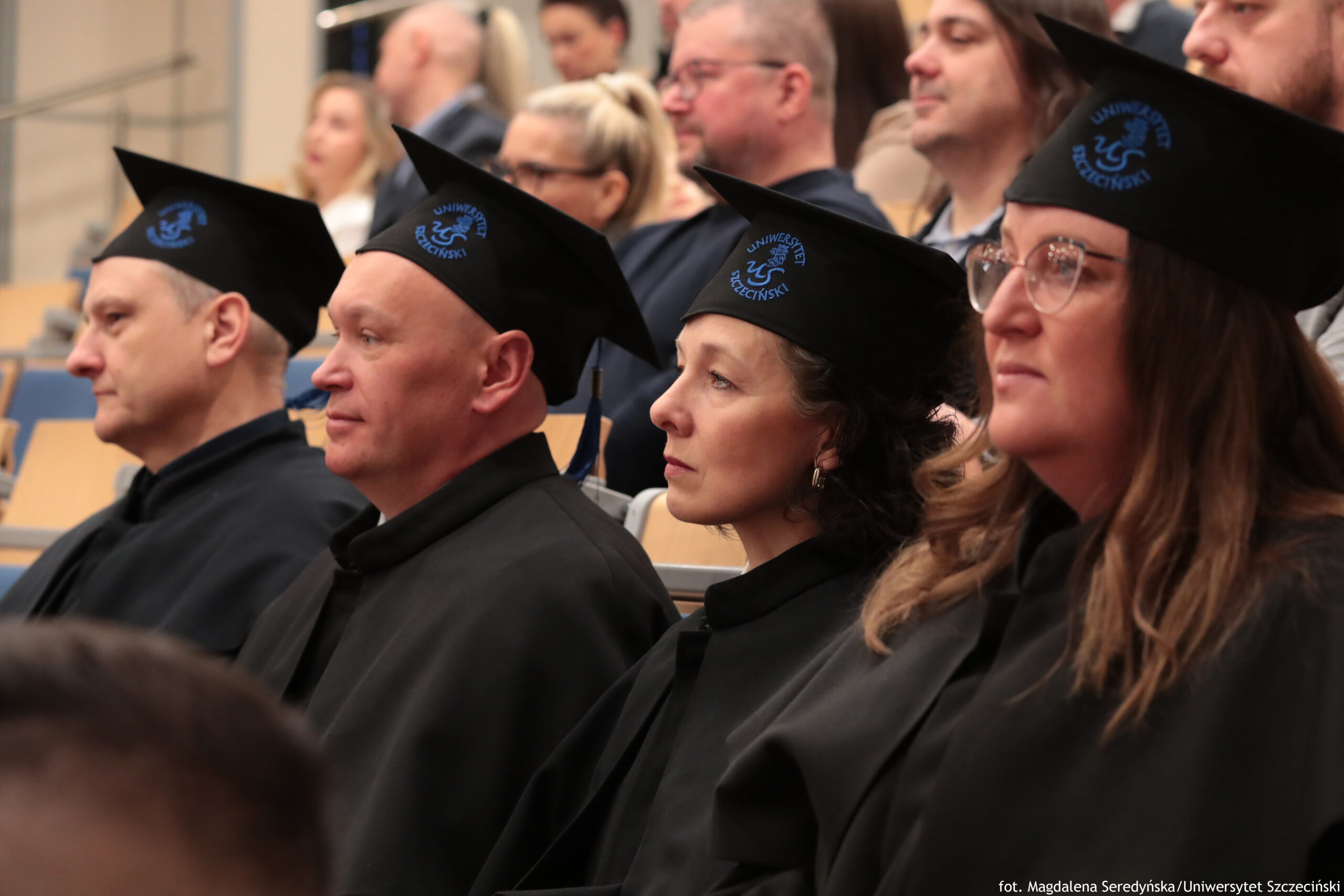 Grupa dorosłych osób w czarnych togach i czarnych biretach akademickich siedząca w auli. Na nakryciach głów widać niebieskie logo i napis „UNIWERSYTET SZCZECIŃSKI”. Na pierwszym planie siedzą cztery osoby, zwrócone bokiem w lewo, z poważnymi, skupionymi wyrazami twarzy. Skóra osób na pierwszym planie jest jasna. Od lewej: mężczyzna z krótkimi włosami; obok mężczyzna łysy; dalej kobieta z ciemnymi, kręconymi włosami do okolic żuchwy; po prawej kobieta w okularach, z długimi brązowymi włosami. W tle, w miękkim rozmyciu, siedzą kolejne osoby na drewnianych ławkach, jak podczas uroczystości akademickiej. W prawym dolnym rogu widoczny jest podpis: „fot. Magdalena Seredyńska/Uniwersytet Szczeciński”.