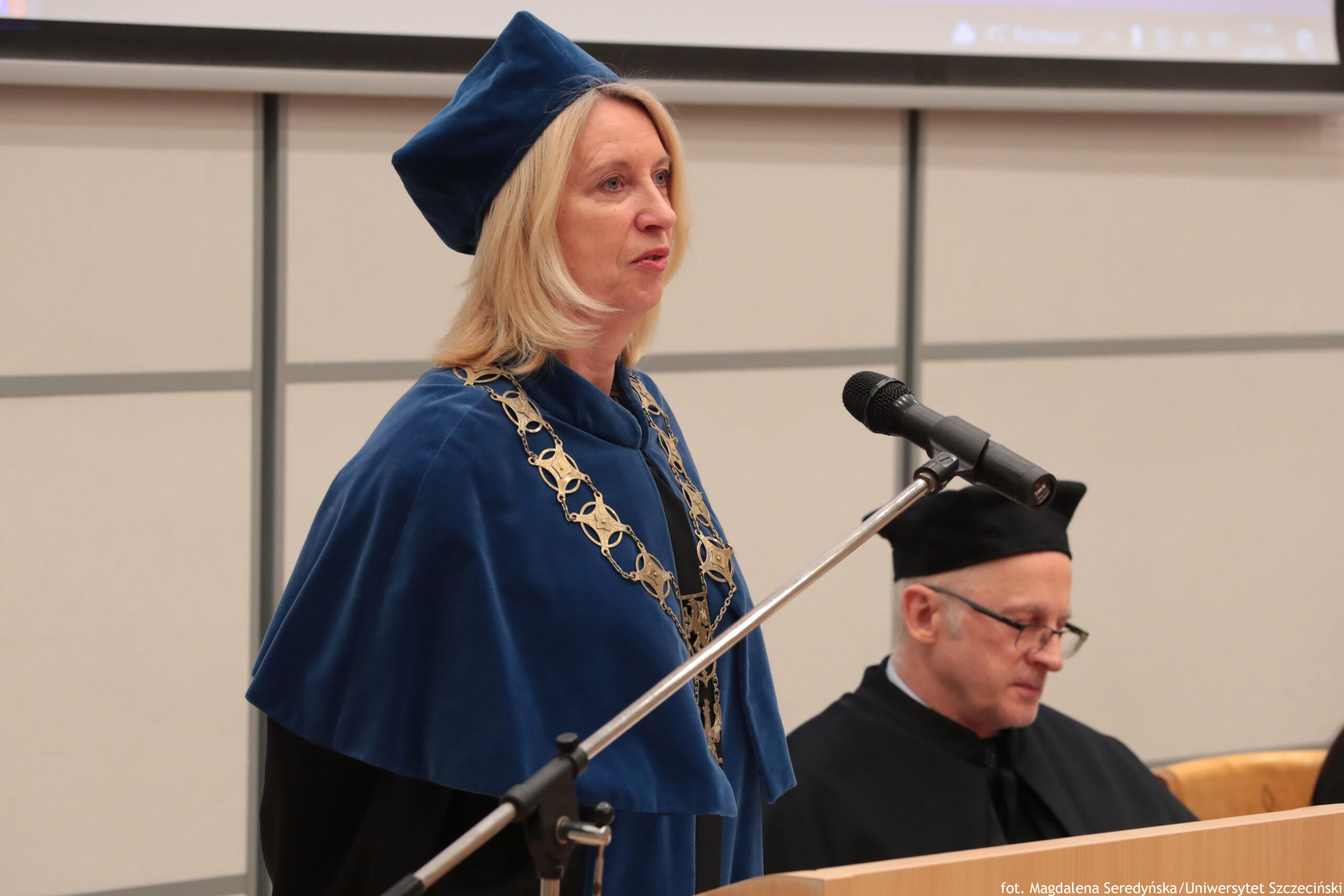 Kobieta w niebieskim stroju akademickim stojąca przy mikrofonie. Ma jasną cerę, blond włosy do ramion i kwadratową niebieską czapkę; na szyi widać ozdobny złoty łańcuch rektorski. Stoi bokiem i wygląda, jakby przemawiała. W tle siedzi mężczyzna w czarnym stroju akademickim i czarnej czapce, w okularach, z opuszczoną głową, jakby czytał lub notował. Na dole zdjęcia widać fragment drewnianej mównicy. W prawym dolnym rogu jest mały podpis: „fot. Magdalena Seredyńska/Uniwersytet Szczeciński”.