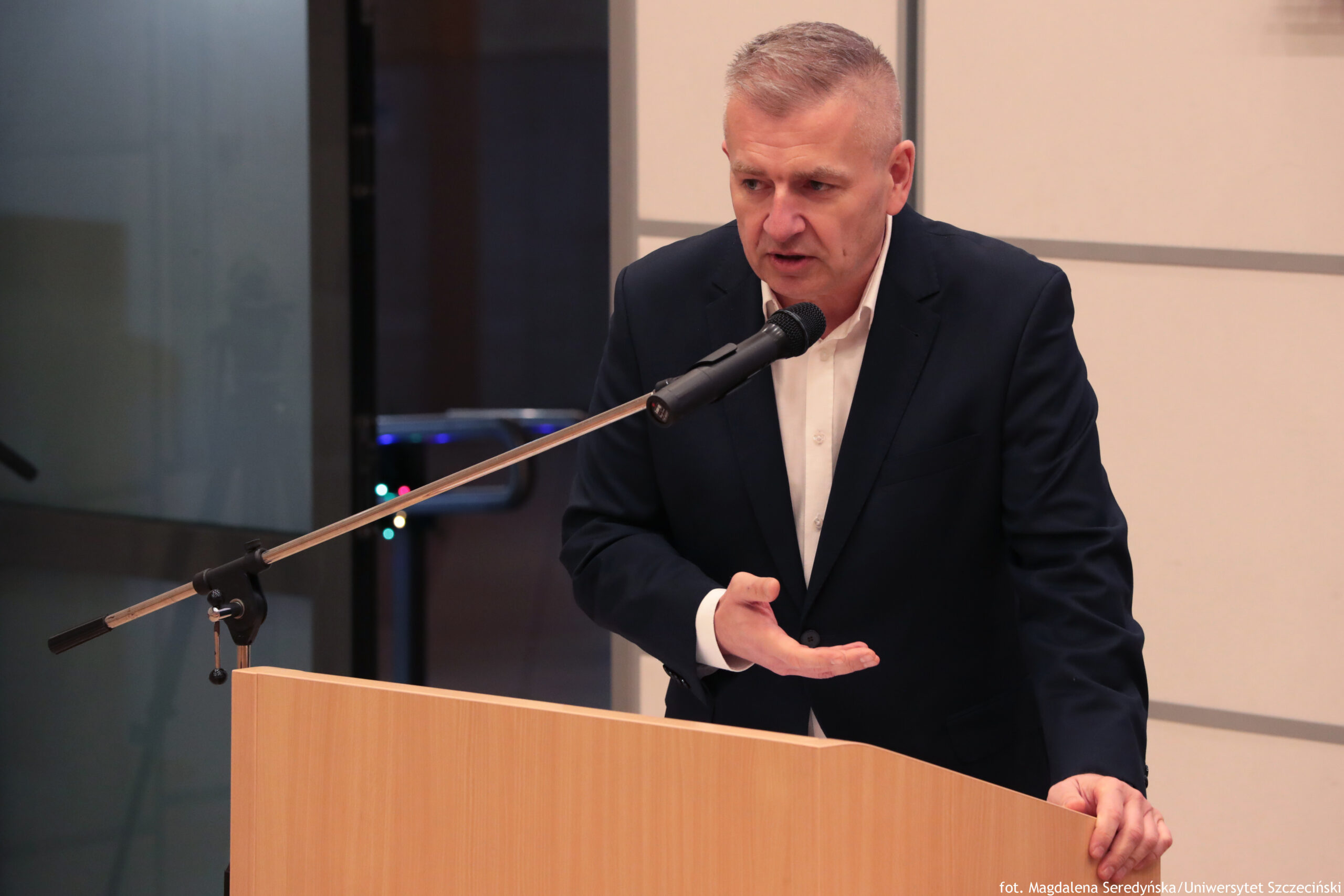 Mężczyzna przemawiający przy drewnianej mównicy do mikrofonu na wysięgniku. Ma jasną skórę, krótkie siwe włosy i poważny wyraz twarzy. Ubrany jest w ciemny garnitur i białą koszulę bez krawata. Jedną dłoń trzyma otwartą na wysokości klatki piersiowej w geście wyjaśniania, drugą opiera na krawędzi mównicy. Tło wygląda na salę konferencyjną z jasną ścianą/panelem oraz ciemniejszą częścią po lewej stronie. W prawym dolnym rogu widoczny jest podpis: „fot. Magdalena Seredyńska / Uniwersytet Szczeciński”.