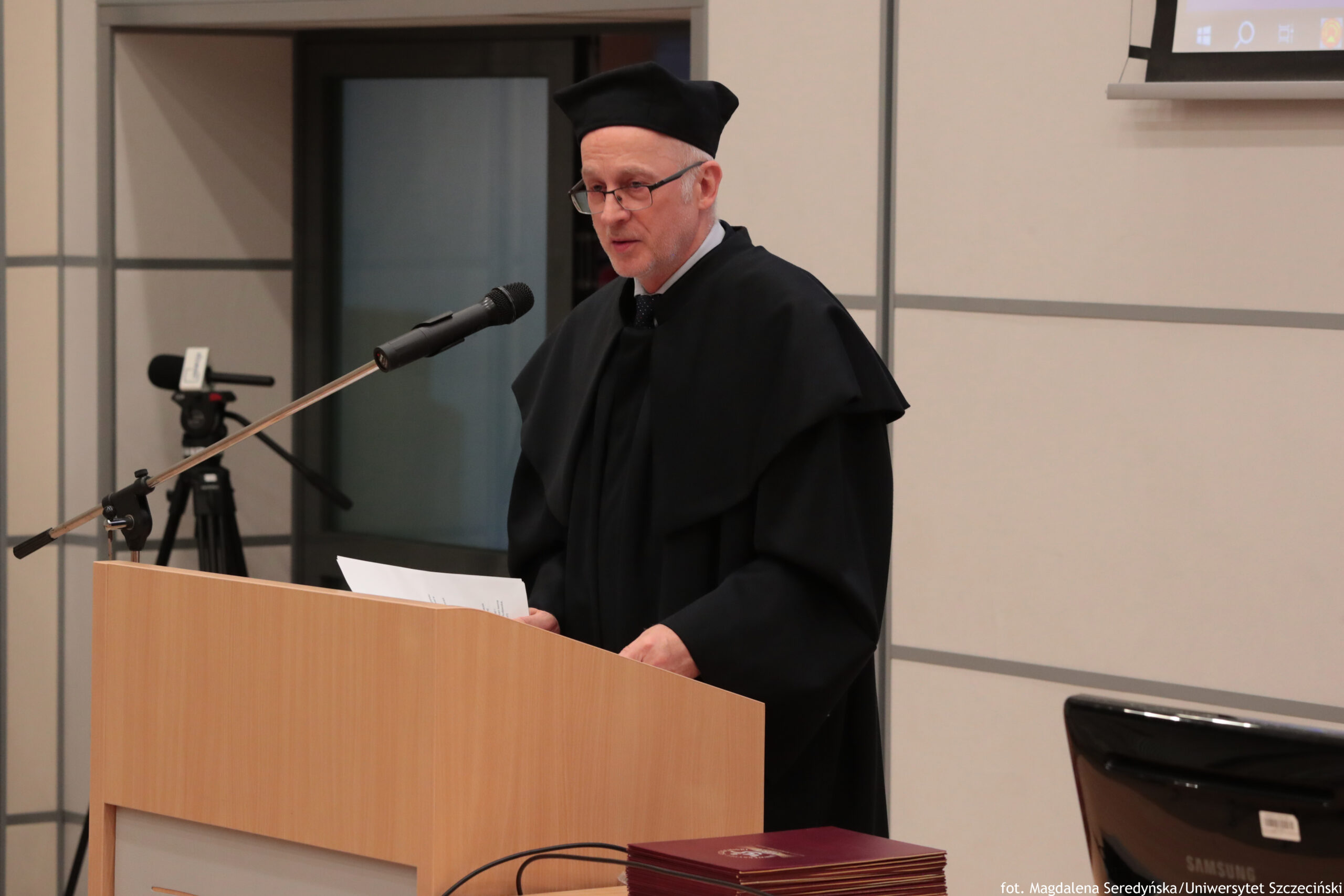 Mężczyzna w czarnej todze przy mównicy z mikrofonem. Ma jasną skórę, okulary, krótko przycięte siwe włosy i czarny biret; trzyma kartki i przemawia. W tle widać wnętrze sali z jasnymi panelami ściennymi, kamerę na statywie po lewej oraz fragment ekranu/projektora u góry po prawej. Na stole z przodu leżą bordowe teczki lub okładki. W prawym dolnym rogu jest drobny podpis fotograficzny: „fot. Magdalena Seredyńska/Uniwersytet Szczeciński”.