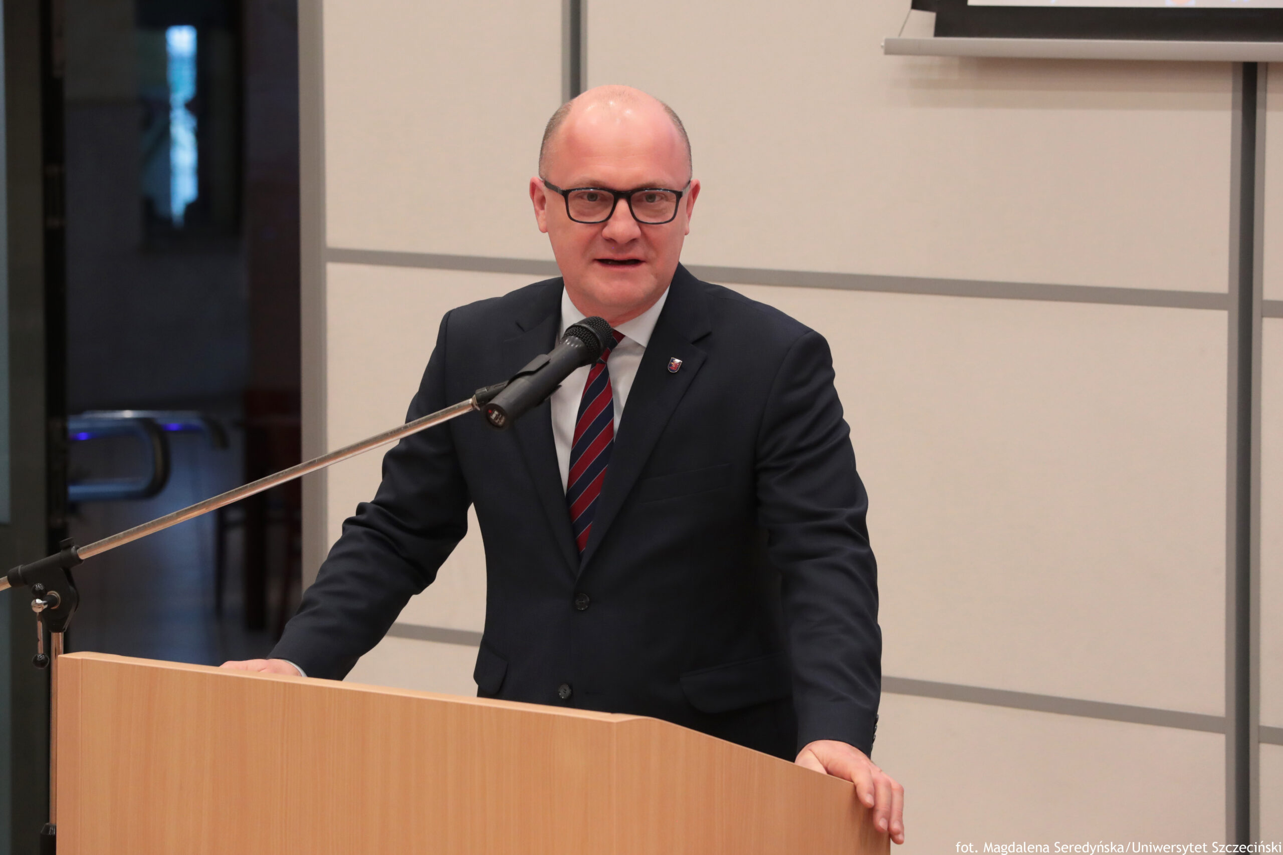 Mężczyzna w garniturze stojący za drewnianą mównicą i przemawiający do mikrofonu. Ma jasną skórę, jest łysy na czubku głowy, nosi czarne okulary. Ubrany jest w ciemny garnitur, białą koszulę i krawat w skośne czerwono-granatowe pasy; na klapie marynarki widać małą przypinkę. Mikrofon na wysięgniku jest ustawiony przed jego ustami. Tło wygląda jak wnętrze sali wykładowej lub konferencyjnej: jasna ściana z poziomymi podziałami, po lewej ciemne drzwi lub przeszklenie, a w prawym górnym rogu fragment ekranu/projektora. W prawym dolnym rogu jest podpis: „fot. Magdalena Seredyńska/Uniwersytet Szczeciński”.
