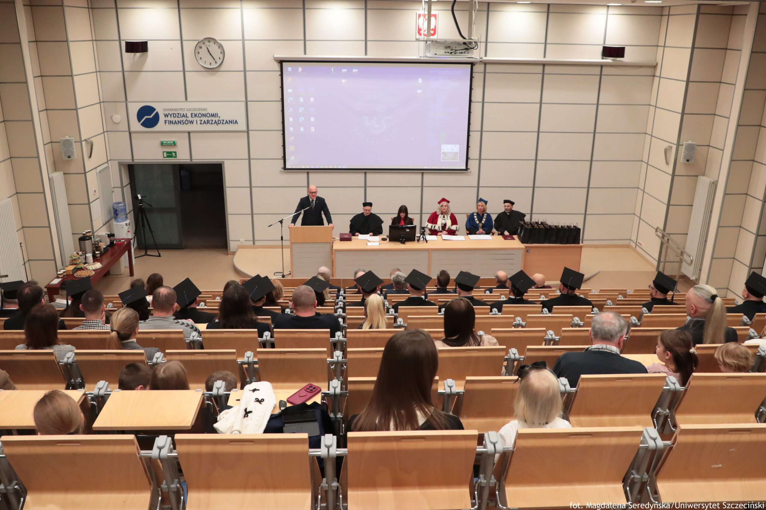 Sala wykładowa z uroczystością akademicką, widok z tyłu na publiczność i scenę z władzami uczelni. W dolnej części kadru widać rzędy drewnianych ławek w auli, zajęte przez wiele osób; część osób ma na głowach czarne birety. Na przodzie sali stoi mównica, przy której przemawia mężczyzna w garniturze. Za nim, przy długim stole, siedzi kilka osób w togach i biretach; jedna osoba ma czerwono-biały strój i łańcuch, inne osoby mają granatowe lub czarne togi i łańcuchy insygniów. Nad sceną wisi duży ekran projekcyjny z wyświetlonym pulpitem komputera. Po lewej stronie na ścianie jest zegar oraz tablica z napisem: „WYDZIAŁ EKONOMII, FINANSÓW I ZARZĄDZANIA”. Po lewej, przy ścianie, stoi stolik z poczęstunkiem. Po prawej stronie sceny ustawiony jest rząd czarnych toreb lub teczek. W prawym dolnym rogu zdjęcia widnieje podpis: „fot. Magdalena Seredyńska/Uniwersytet Szczeciński”.