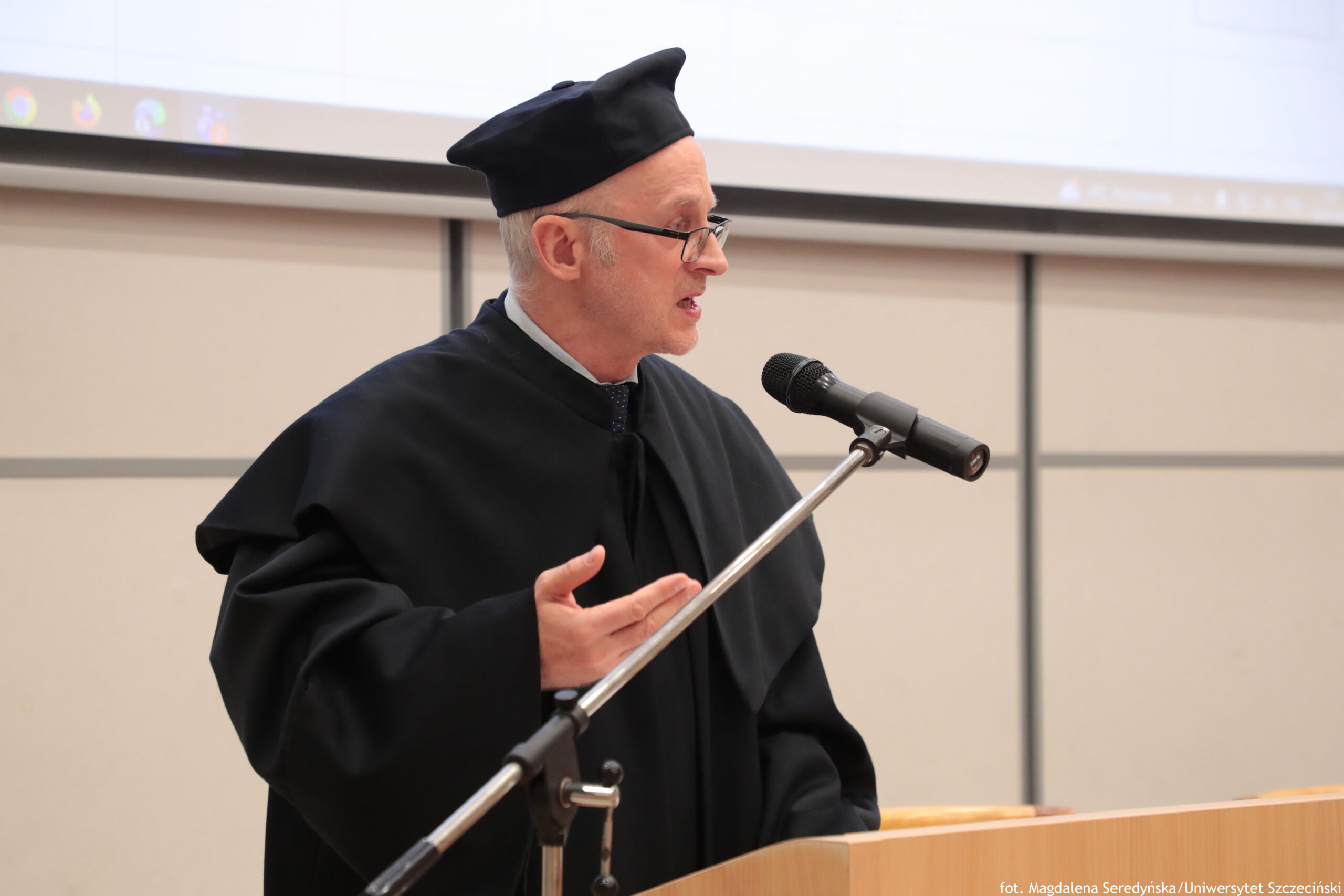 Mężczyzna w czarnym stroju akademickim i czarnym birecie, przemawiający do mikrofonu. Ma jasną skórę, krótkie siwe włosy po bokach i okulary; jest pokazany z profilu, z otwartymi ustami w trakcie mówienia i uniesioną dłonią w geście. Stoi przy drewnianej mównicy, a mikrofon jest na metalowym statywie. W tle widać jasną ścianę a u góry fragment ekranu/projektora. W prawym dolnym rogu jest mały napis: „fot. Magdalena Seredyńska/Uniwersytet Szczeciński”.