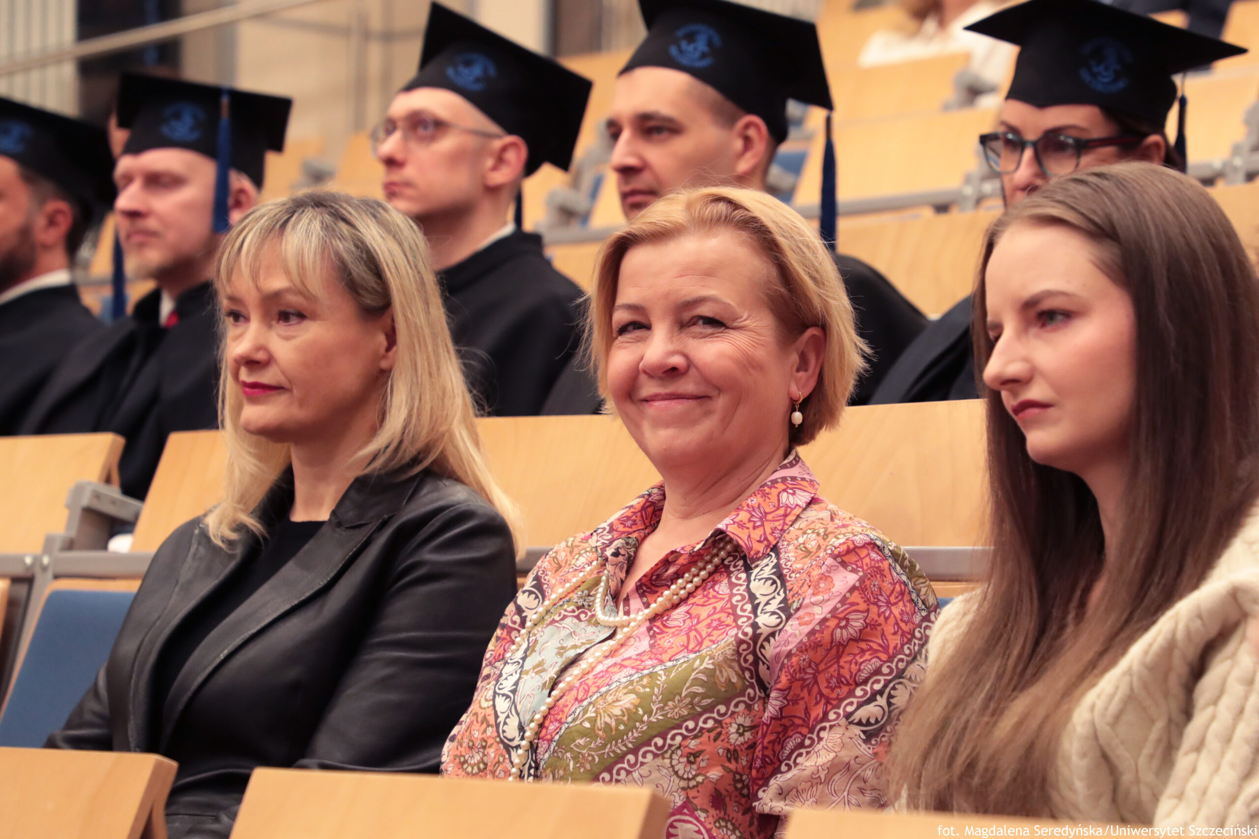 Grupa osób siedzących w auli, część w czarnych togach i biretach. W pierwszym rzędzie siedzą trzy kobiety. Po lewej kobieta o jasnej cerze i blond włosach do ramion z grzywką, w czarnej skórzanej kurtce, z czerwonymi ustami; ma poważny wyraz twarzy i patrzy w bok. Na środku kobieta o jasnej cerze i krótkich blond włosach, uśmiechnięta; ma kolczyki i kolorową, wzorzystą bluzkę w odcieniach różu i pomarańczu oraz naszyjnik z jasnych korali. Po prawej młoda kobieta o jasnej cerze i długich, prostych brązowych włosach, w jasnym swetrze; ma neutralny, lekko zamyślony wyraz twarzy i patrzy w bok. W tle siedzą mężczyźni w czarnych togach i czapkach absolwenckich, niektórzy w okularach. Na dole po prawej widoczny jest napis: „fot. Magdalena Seredyńska / Uniwersytet Szczeciński”.