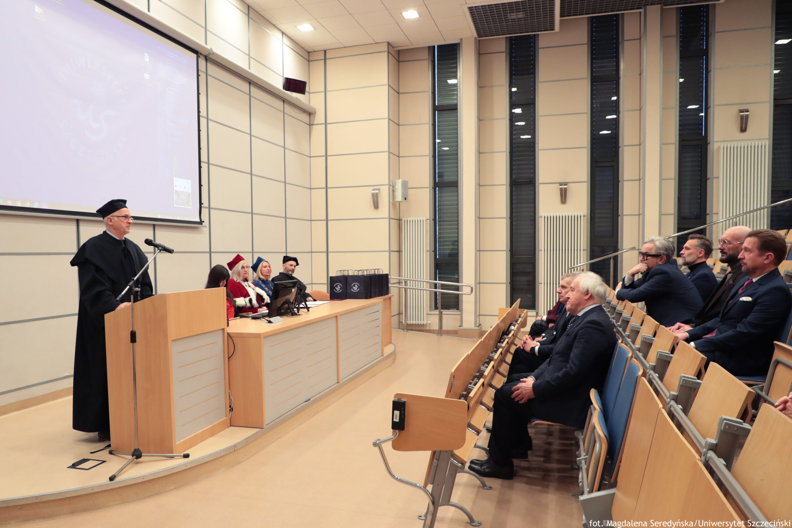 Sala wykładowa z uroczystością akademicką, z mównicą po lewej i publicznością po prawej. Po lewej stronie stoi starszy mężczyzna w czarnej todze i birecie, przemawia do mikrofonu przy drewnianej mównicy. Za nim, przy długim stole, siedzi kilka osób w strojach akademickich; część ma czerwone elementy (czapki i peleryny), jedna osoba ma niebieski akcent. Na stole stoją ciemne torby lub teczki z jasnym logo. Po prawej stronie widać rzędy drewnianych krzeseł z niebieskimi siedziskami i grupę mężczyzn w garniturach siedzących w pierwszych rzędach, zwróconych w stronę mówcy. W tle są wysokie, wąskie okna oraz jasne ściany z panelami. W lewym górnym rogu jest ekran projekcyjny z niewyraźnym obrazem. W prawym dolnym rogu widoczny podpis: „fot. Magdalena Seredyńska / Uniwersytet Szczeciński”.