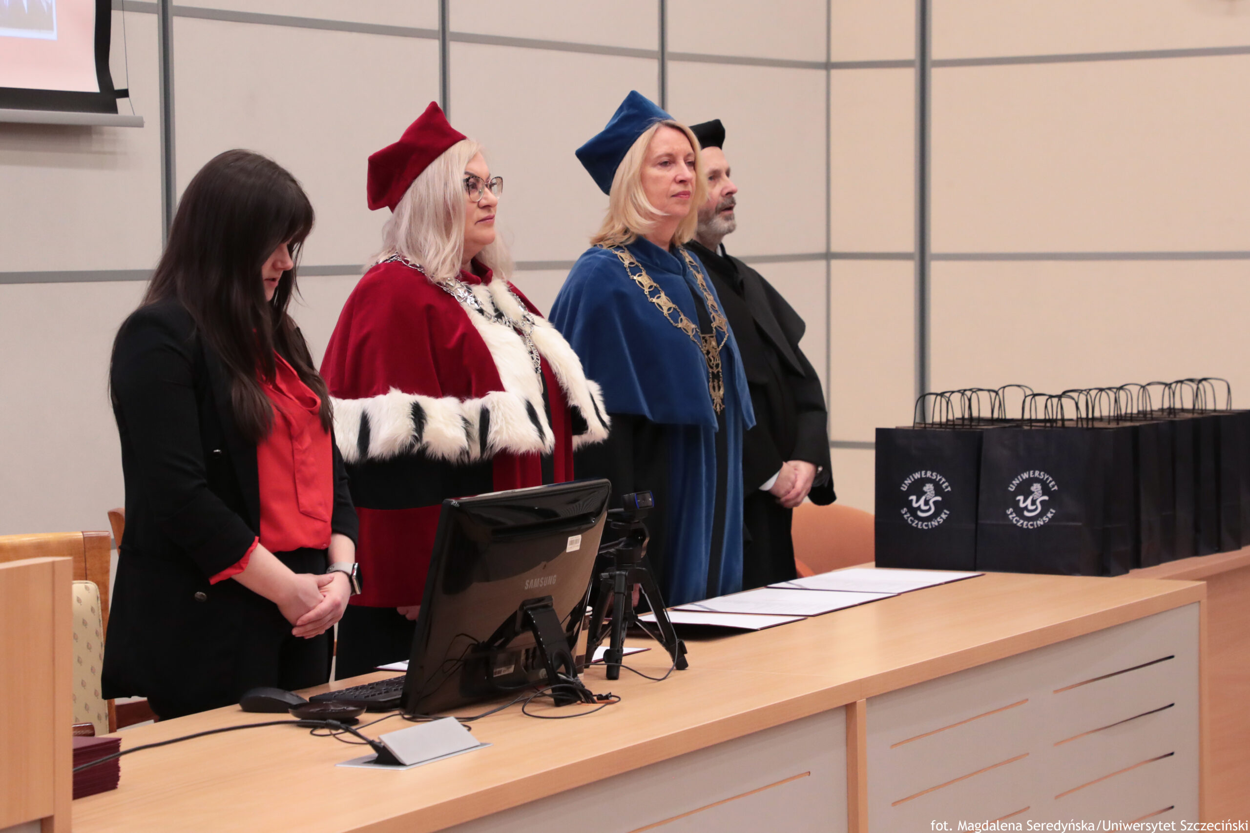 Grupa czterech osób stojących przy długim stole w sali wykładowej, dwie osoby w uroczystych strojach akademickich. Po lewej stoi kobieta o jasnej skórze z długimi, ciemnymi włosami, w czarnej marynarce i czerwonej bluzce, z głową opuszczoną i rękami złożonymi z przodu. Obok niej stoi kobieta o jasnej skórze z siwymi włosami i okularami, w czerwonej todze z białym futrzanym wykończeniem oraz czerwonym biretem. Dalej stoi kobieta o jasnej skórze z blond włosami, w niebieskiej todze i niebieskim birecie, z ozdobnym łańcuchem na szyi. Za nią częściowo widoczny jest mężczyzna o jasnej skórze z siwą brodą, w czarnym stroju i czarnym nakryciu głowy. Na stole stoją monitor komputerowy i akcesoria, leżą też dokumenty. Po prawej stronie na stole ustawiono kilka czarnych toreb prezentowych z białym logo i napisem „Uniwersytet Szczeciński” oraz „US”. W prawym dolnym rogu widoczny jest podpis: „fot. Magdalena Seredyńska/Uniwersytet Szczeciński”.