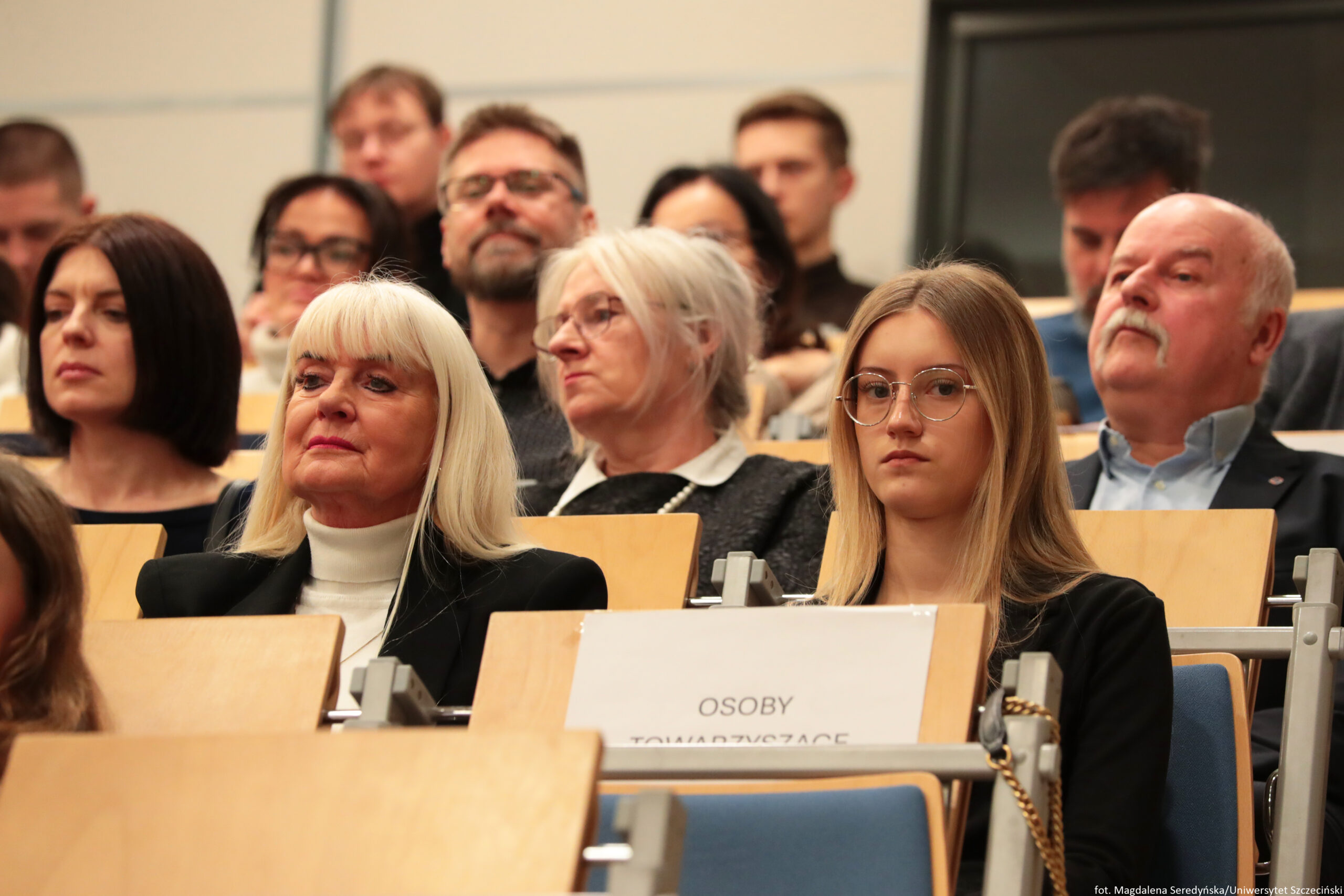 Grupa dorosłych osób siedzących na sali wykładowej lub konferencyjnej, w rzędach z drewnianymi pulpitami. W pierwszym rzędzie na środku siedzi starsza kobieta o jasnej cerze z prostymi, długimi, bardzo jasnymi blond włosami i grzywką, w czarnym żakiecie i białym golfie. Obok niej, po prawej stronie kadru, siedzi młoda kobieta o jasnej cerze z długimi blond włosami i okrągłymi okularami, w ciemnym ubraniu; ma neutralny, skupiony wyraz twarzy. Po lewej stronie pierwszego rzędu widać kobietę o jasnej cerze z ciemnym, prostym bobem, także w ciemnym stroju, patrzącą przed siebie. W tle siedzą kolejne osoby, m.in. starsza kobieta o siwych włosach w okularach oraz starszy mężczyzna o jasnej cerze z siwymi włosami i wyraźnym wąsem, w jasnej koszuli. Na pulpicie przed młodą kobietą znajduje się kartka z napisem „OSOBY TOWARZYSZĄCE”. W prawym dolnym rogu widnieje mały podpis: „fot. Magdalena Seredyńska/Uniwersytet Szczeciński”.