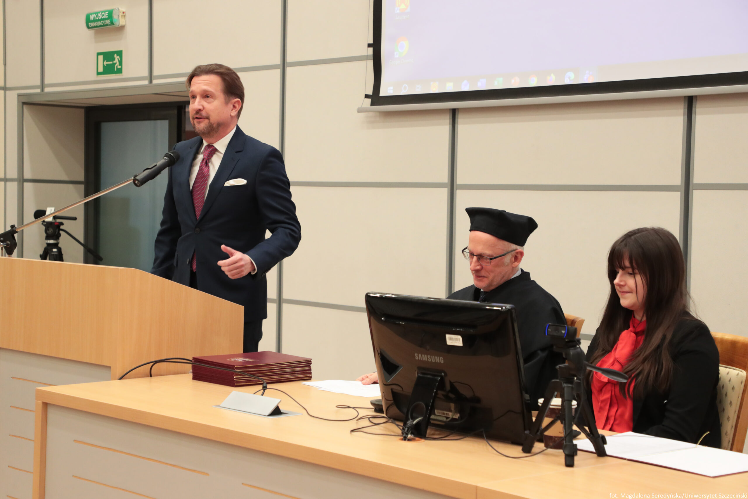 Mężczyzna w granatowym garniturze, białej koszuli i bordowym krawacie przemawia przy mównicy do mikrofonu w sali wykładowej. Ma krótkie brązowe włosy i jasną skórę. Po prawej stronie przy długim stole siedzą dwie osoby: starszy mężczyzna w czarnym stroju i czarnym birecie oraz kobieta z długimi ciemnymi włosami, w czarnym ubraniu z czerwonym szalikiem. Na stole stoi monitor komputerowy Samsung, leżą dokumenty i ułożony stos czerwonych teczek. Z przodu stołu widać też mały statyw. W tle są jasne, panelowe ściany, fragment opuszczonego ekranu projekcyjnego oraz zielony znak „WYJŚCIE” i piktogram wyjścia ewakuacyjnego. W prawym dolnym rogu znajduje się drobny napis, nieczytelny w tej jakości.