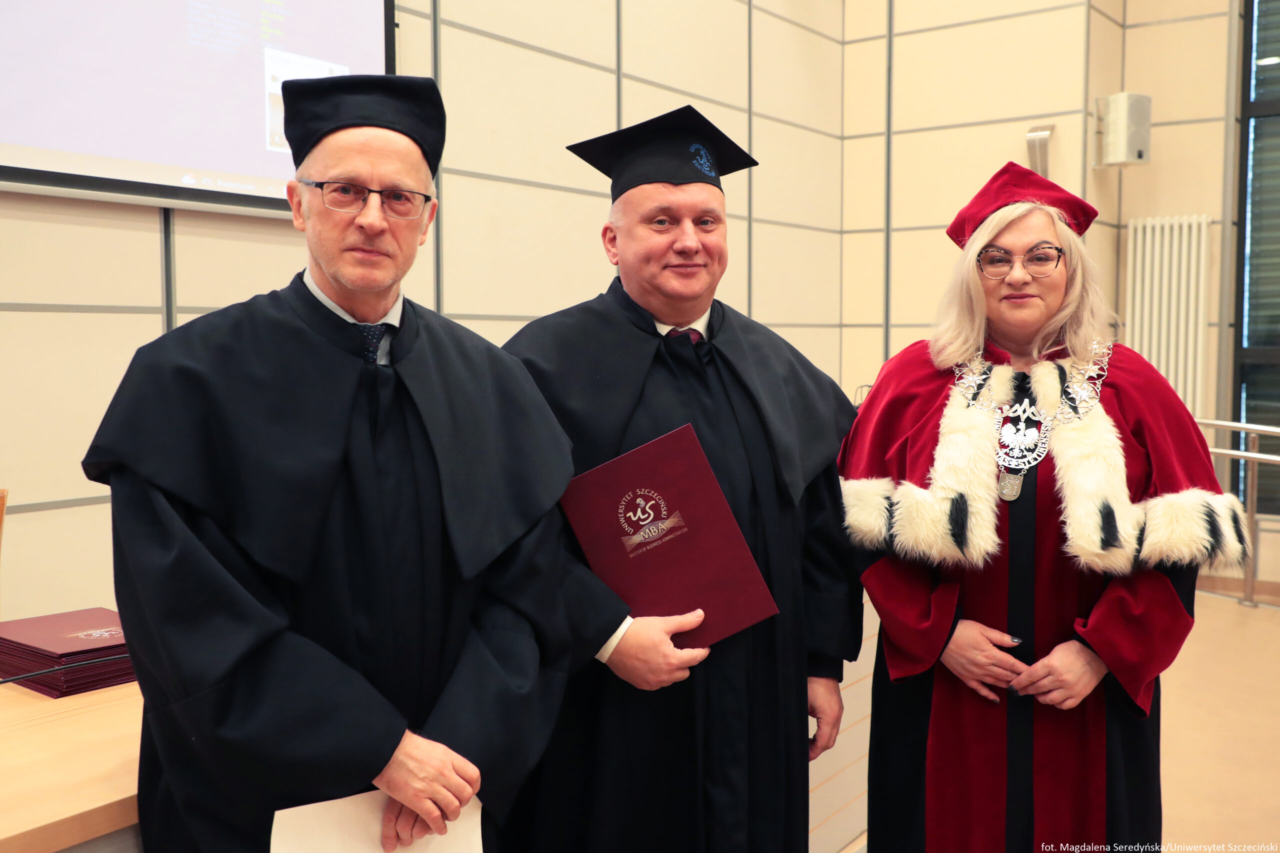 Trzy osoby w akademickich togach stojące obok siebie w sali. To zdjęcie wygląda na uroczystość uczelnianą. Po lewej stoi mężczyzna o jasnej skórze w czarnej todze i czarnym, płaskim nakryciu głowy; ma okulary i poważny wyraz twarzy, w rękach trzyma jasny dokument. W środku stoi mężczyzna o jasnej skórze w czarnej todze i czarnym birecie; lekko się uśmiecha i trzyma bordową teczkę/dyplom z widocznym białym godłem/logo i napisem „Uniwersytet Szczeciński” oraz „MBA”. Po prawej stoi kobieta o jasnej skórze w czerwonej todze z białym futrzanym obszyciem i czarnymi wstawkami; ma jasne blond włosy, okulary i na szyi duży ozdobny łańcuch z medalionem z orłem. W tle widać jasne ściany z geometrycznymi podziałami, fragment ekranu/projektora, stół z ułożonymi bordowymi teczkami oraz pionowy grzejnik. W prawym dolnym rogu jest drobny podpis: „fot. Magdalena Seredyńska/Uniwersytet Szczeciński”.
