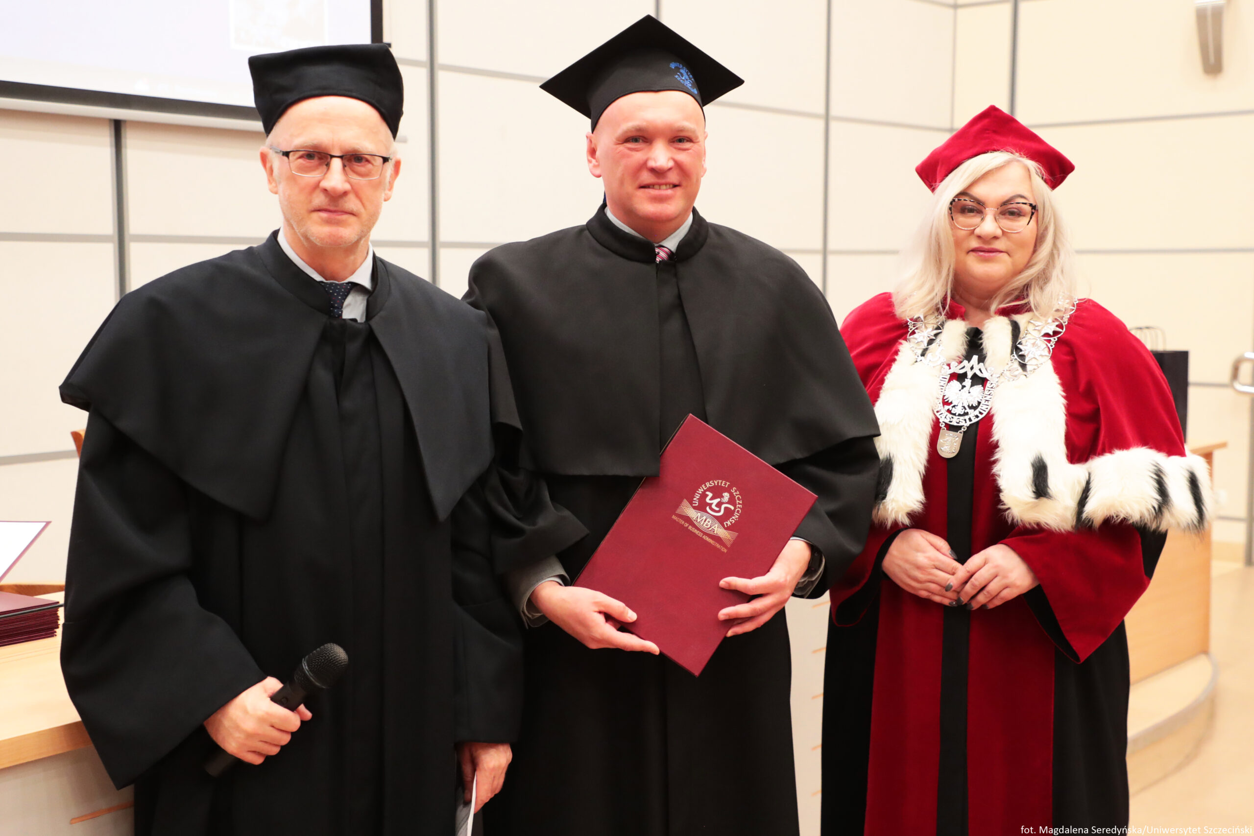 Trzy osoby w strojach akademickich podczas uroczystości na uczelni. Po lewej stoi mężczyzna w czarnej todze i czarnym birecie, w okularach, trzyma mikrofon. W środku stoi mężczyzna w czarnej todze i czarnym birecie, uśmiecha się i trzyma bordową teczkę z godłem oraz napisem „UNIWERSYTET SZCZECIŃSKI” i „MBA”. Po prawej stoi kobieta w czerwonej todze z białym futrzanym obszyciem i czerwonym birecie, w okularach, ma na szyi ozdobny łańcuch z medalionem. Tło to jasna sala wykładowa z podestem i ścianą z paneli. W prawym dolnym rogu jest podpis: „fot. Magdalena Seredyńska/Uniwersytet Szczeciński”.