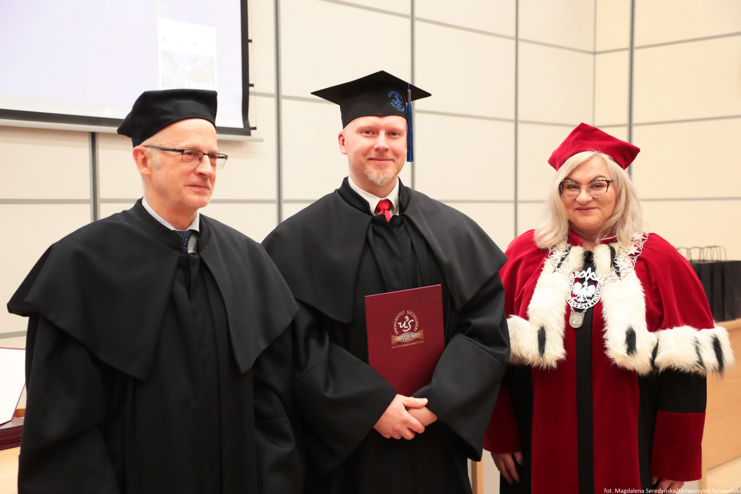 Trzy osoby w strojach akademickich podczas uroczystości. Po lewej stoi mężczyzna o jasnej skórze w czarnej todze i czarnym birecie, w okularach, zwrócony lekko w stronę środka. W środku stoi mężczyzna o jasnej skórze w czarnej todze i czarnym birecie z niebieskim chwostem; ma jasny zarost i trzyma przed sobą bordowy dyplom lub teczkę z jasnym godłem i skrótem „MBA”. Po prawej stoi kobieta o jasnej skórze w czerwonej todze z białym futrzanym obszyciem i czerwonym birecie; ma jasne, długie włosy, okulary i na szyi duży srebrny łańcuch z medalionem. Tło to jasna ściana z podziałem na prostokątne panele oraz fragment ekranu projekcyjnego po lewej. W prawym dolnym rogu widoczny jest mały podpis: „fot. Magdalena Seredyńska/Uniwersytet Szczeciński”.
