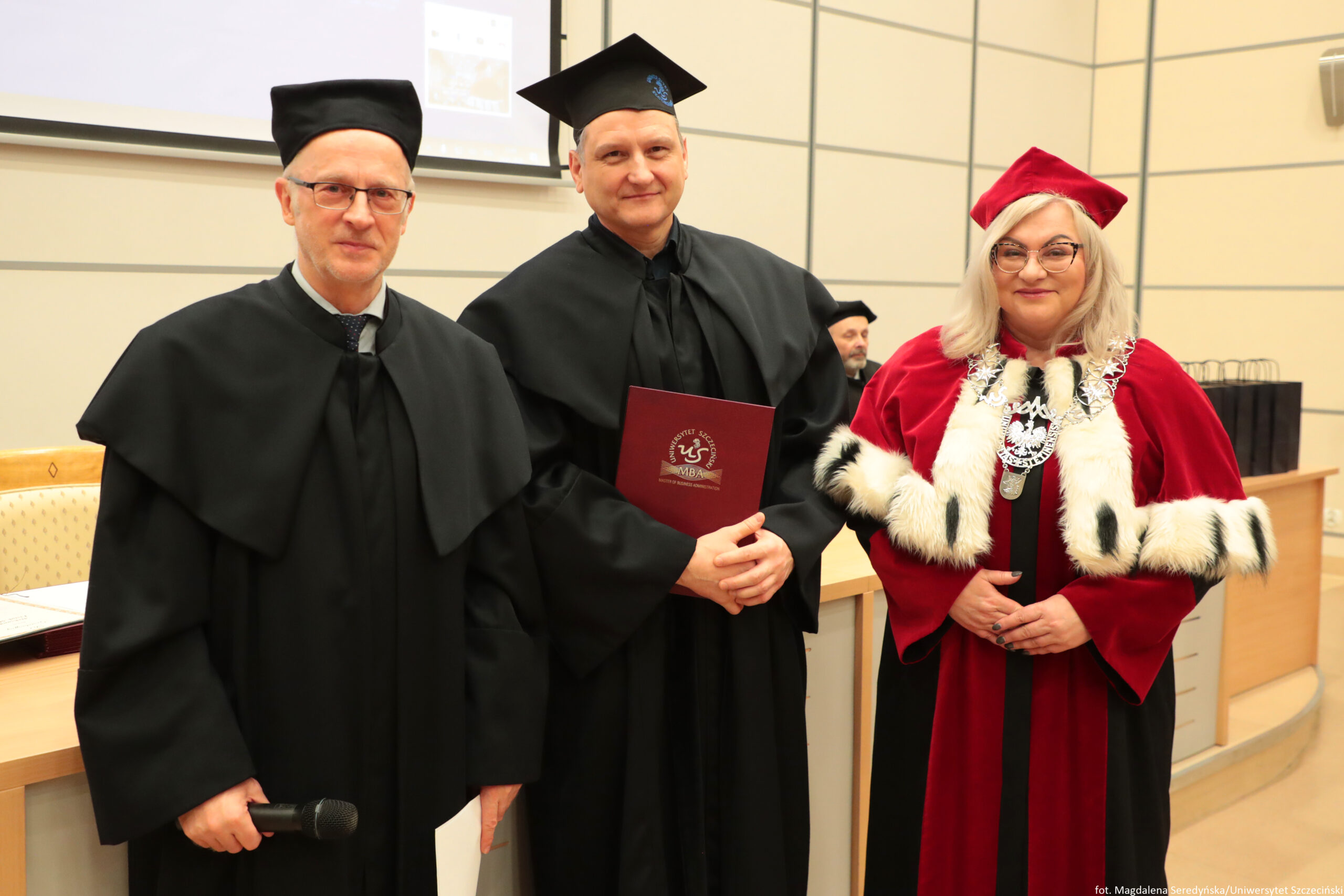 Trzy osoby w akademickich strojach stojące w sali wykładowej. Po lewej stronie stoi mężczyzna w czarnej todze i czarnym birecie; ma jasną skórę, okulary i trzyma w dłoni mikrofon. W środku stoi mężczyzna w czarnej todze i czarnym birecie; ma jasną skórę i trzyma bordową teczkę z białym godłem i napisem „MBA”. Po prawej stronie stoi kobieta w czerwonej todze i czerwonym birecie; ma jasną skórę, jasne blond włosy, okulary oraz bogaty łańcuch rektorski na szyi, a toga ma białe futrzane wykończenia z czarnymi plamkami. W tle widać fragment ekranu/projektora oraz elementy sali, a także siedzącą, częściowo zasłoniętą osobę. W prawym dolnym rogu jest drobny podpis fotograficzny: „fot. Magdalena Seredyńska/Uniwersytet Szczeciński”.