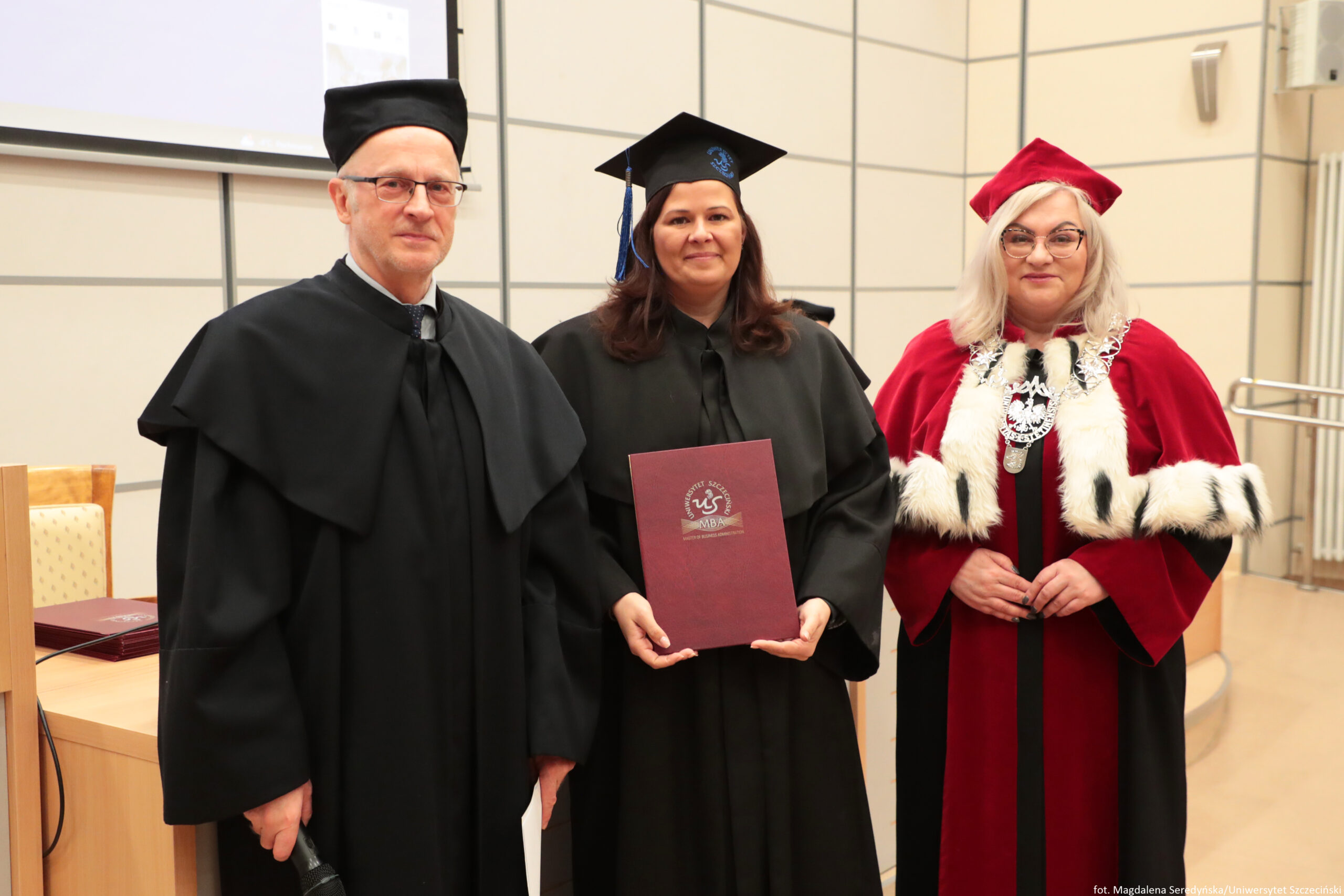 Trzy osoby w strojach akademickich stoją obok siebie w sali, a środkowa osoba trzyma bordową teczkę z dyplomem. Po lewej stoi starszy mężczyzna o jasnej skórze, w czarnym todze i czarnym birecie, w okularach. W środku stoi kobieta o jasnej skórze, w czarnej todze i czarnym birecie z niebieskim chwostem; trzyma przed sobą bordową teczkę z widocznym logo i napisem „MBA”. Po prawej stoi kobieta o jasnej skórze, w czerwonej todze z białym futrzanym wykończeniem i czerwonym birecie; ma jasne, blond włosy i okulary oraz nosi ozdobny łańcuch rektorski. W tle widać jasne ściany z podziałami na panele, fragment ekranu/projektora oraz elementy sali wykładowej. W prawym dolnym rogu jest drobny podpis fotograficzny, ale jest zbyt mały, by go pewnie odczytać.