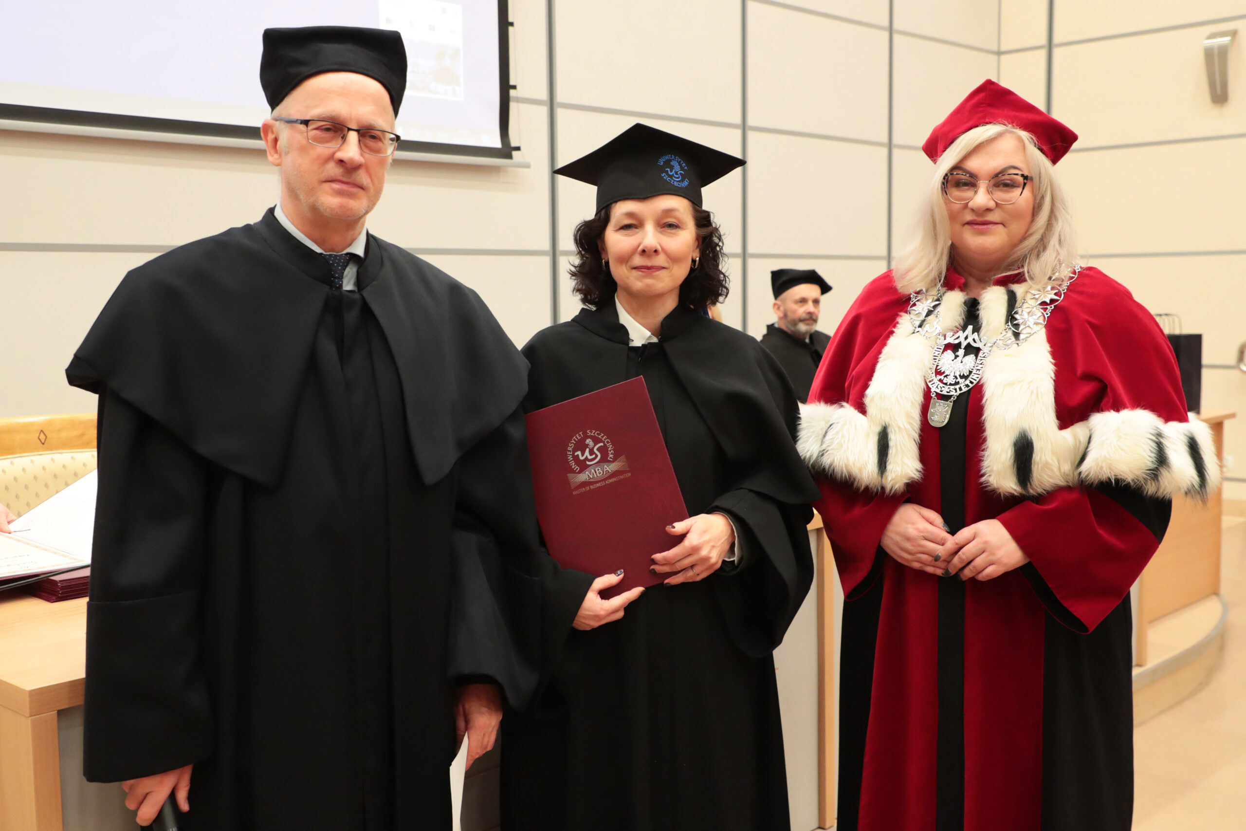 Trzy osoby w akademickich togach podczas uroczystości na sali wykładowej. Pośrodku stoi kobieta w czarnej todze i birecie, trzyma bordową teczkę z napisem „Uniwersytet Śląski w Katowicach” oraz skrótem „MBA”. Po lewej stoi mężczyzna w czarnej todze i czapce, ma okulary. Po prawej stoi kobieta w czerwonej todze z białym futrzanym obszyciem i czerwonym biretem, ma okulary i ozdobny łańcuch rektorski na piersi. W tle widać fragment ekranu projekcyjnego i jeszcze jedną osobę w czarnym stroju akademickim.