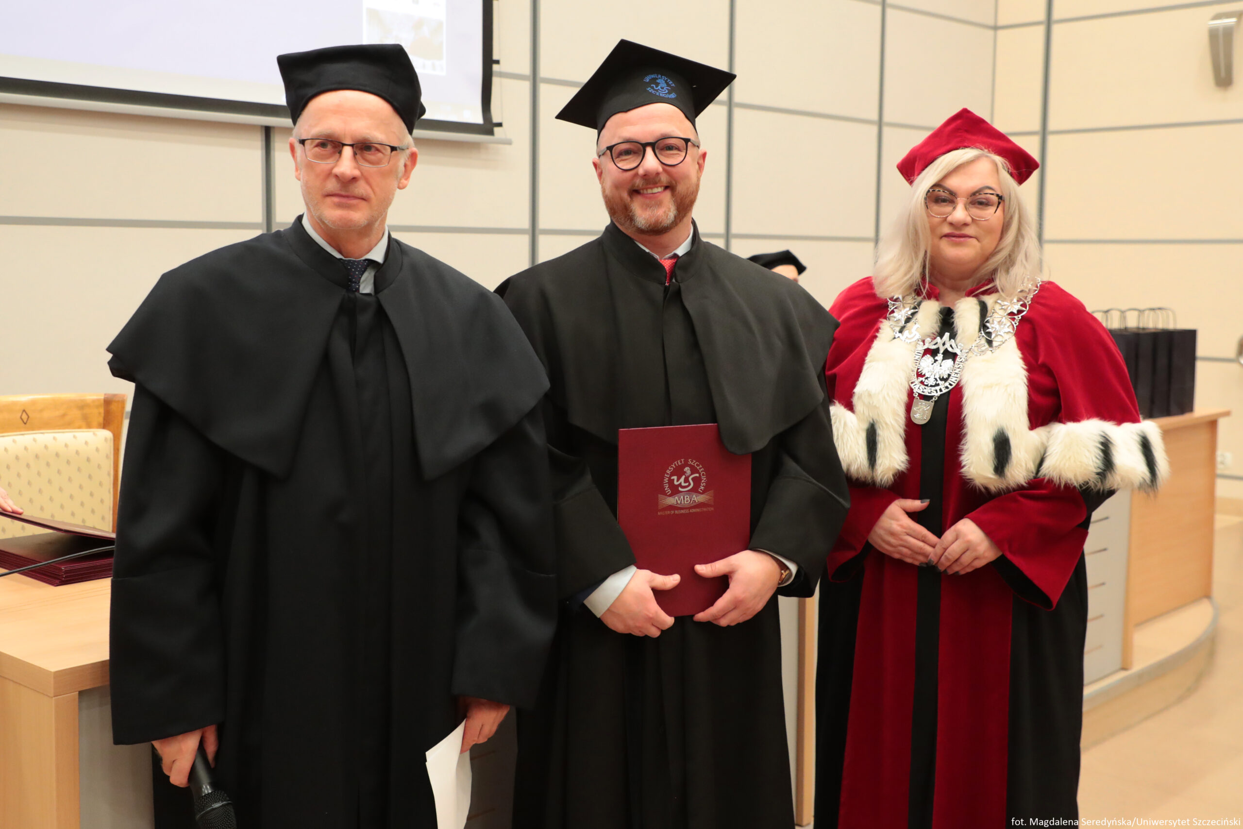 Trzy osoby w strojach akademickich stojące obok siebie w sali, prawdopodobnie podczas uroczystości na uczelni. Po lewej stronie stoi mężczyzna o jasnej karnacji w czarnej todze i czarnym birecie, w okularach; ma poważny wyraz twarzy. Na środku stoi mężczyzna o jasnej karnacji w czarnej todze i czarnym birecie, w okularach, z krótką brodą; uśmiecha się i trzyma bordową teczkę/dyplom z widocznym białym godłem i napisem „MBA”. Po prawej stronie stoi kobieta o jasnej karnacji w czerwonym stroju akademickim z białym futrzanym wykończeniem przy rękawach i przodzie, w czerwonym birecie i okularach; na szyi ma duży, ozdobny łańcuch z medalionem. W tle widać jasne ściany z podziałem na kwadratowe panele, fragment ekranu/projektora u góry oraz biurko i krzesło po lewej. W prawym dolnym rogu jest drobny napis: „fot. Magdalena Seredyńska/Uniwersytet Szczeciński”.