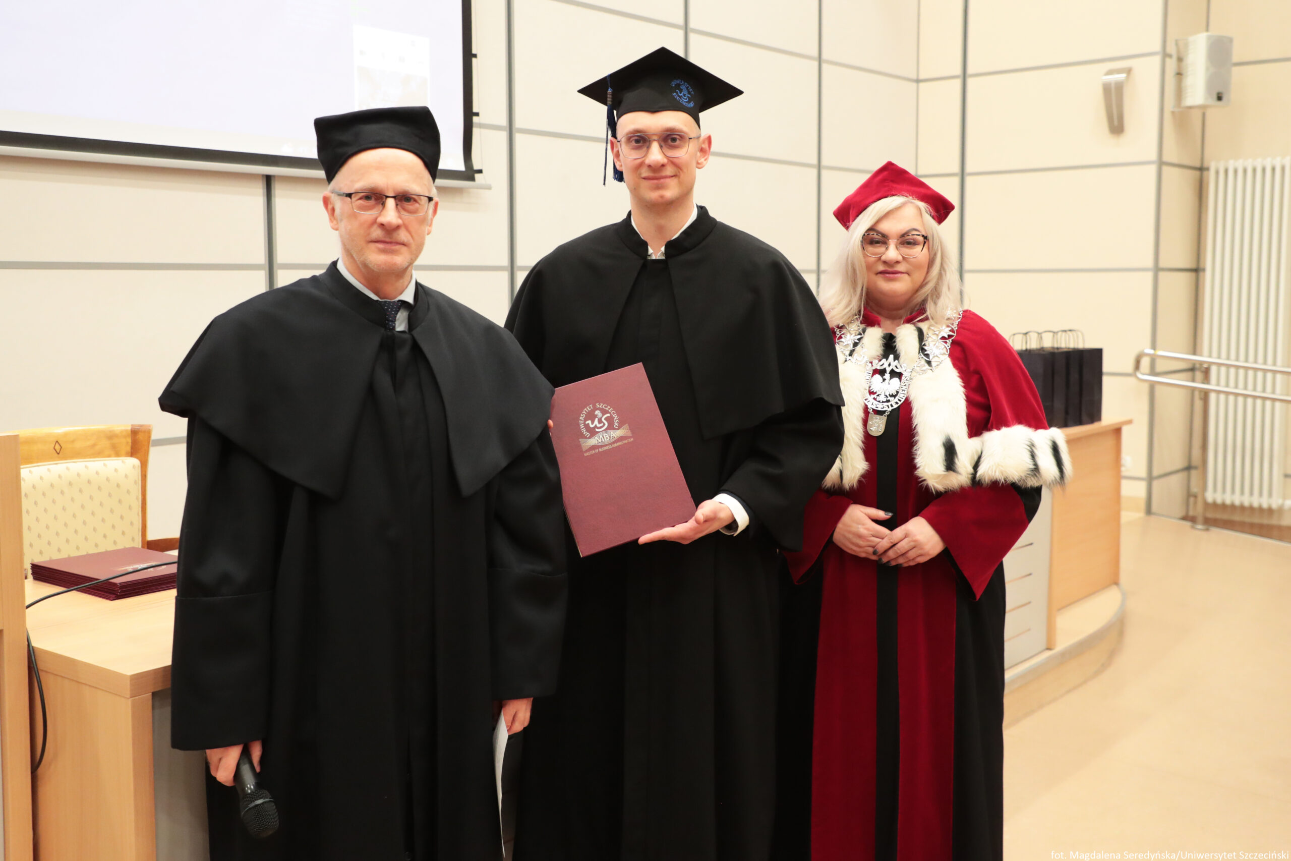 Trzy osoby w strojach akademickich pozujące do zdjęcia w sali uczelni, podczas uroczystości. Środkowa osoba w czarnej todze i birecie trzyma bordowy dyplom z godłem i napisem „Uniwersytet Szczeciński”. Po lewej stoi mężczyzna w czarnej todze i czarnym nakryciu głowy, w okularach, trzyma mikrofon. Po prawej stoi kobieta w czerwonej todze z białym futrzanym wykończeniem i czerwonym nakryciem głowy, w okularach, z dużym łańcuchem insygniów na piersi. W tle widać jasne ściany z panelami, biurko, krzesło oraz fragment ekranu projekcyjnego. W prawym dolnym rogu jest mały podpis: „fot. Magdalena Smoczyńska/Uniwersytet Szczeciński”.”