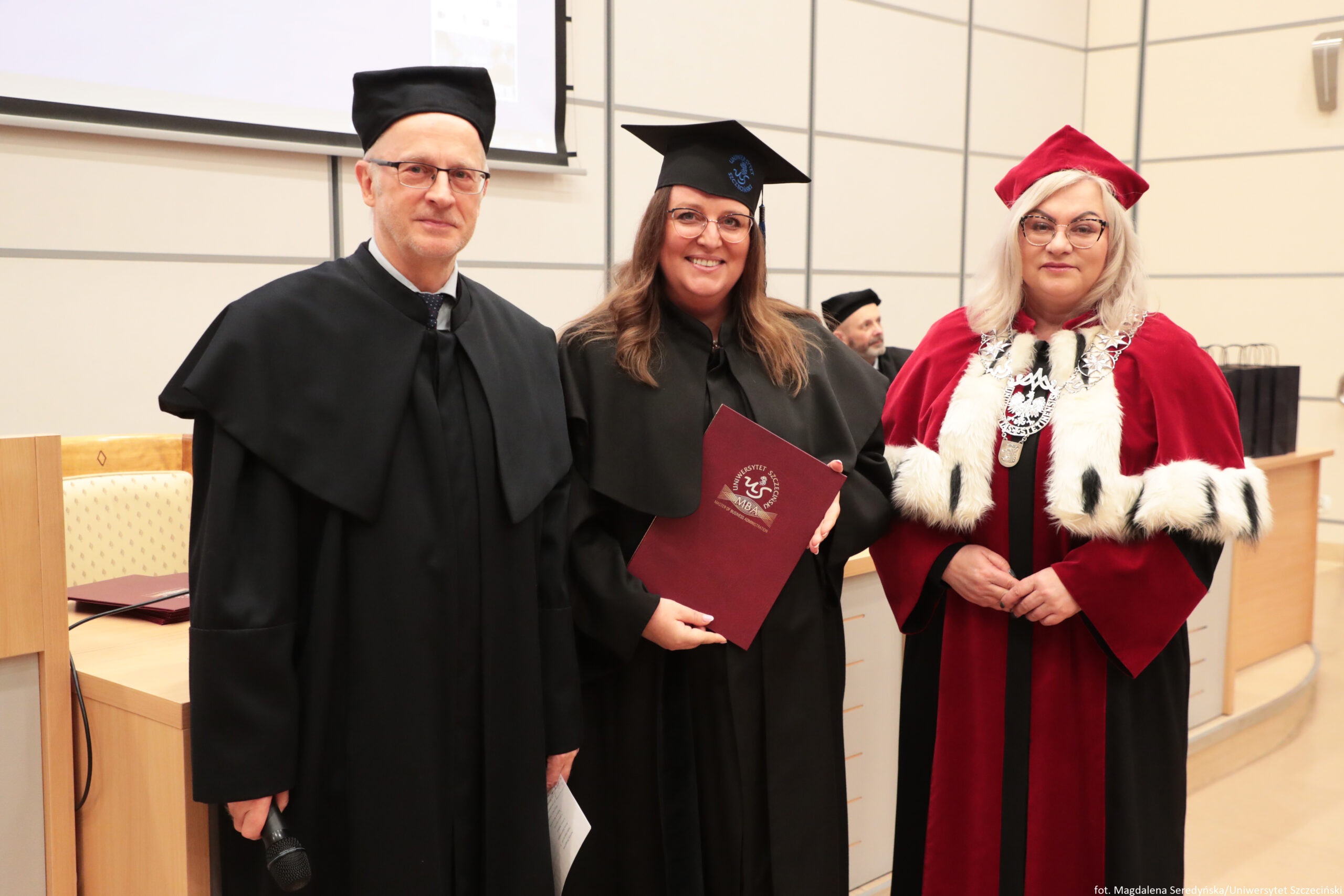 Trzy osoby w strojach akademickich stojące obok siebie w sali wykładowej. Na pierwszym planie widać mężczyznę po lewej, kobietę pośrodku i kobietę po prawej. Mężczyzna po lewej ma jasną skórę, okulary, krótko ostrzyżone włosy i czarny strój akademicki z czarną czapką. W dłoni trzyma mikrofon. Kobieta pośrodku ma jasną skórę, długie brązowe włosy, okulary i czarną togę oraz czarny biret. Uśmiecha się i trzyma bordową teczkę/dyplom z białym godłem i napisem. Kobieta po prawej ma jasną skórę, jasne blond włosy do ramion, okulary i czerwony strój ceremonialny z białym futrzanym wykończeniem oraz czerwony biret. Na piersi ma duży ozdobny łańcuch z medalionem. W tle są jasne ściany z podziałami, fragment ekranu/projektora oraz drewniane biurko i krzesła; widać też siedzącą, rozmytą postać w czapce akademickiej. W prawym dolnym rogu jest mały podpis „fot. Magdalena Seredyńska/Uniwersytet Szczeciński”.