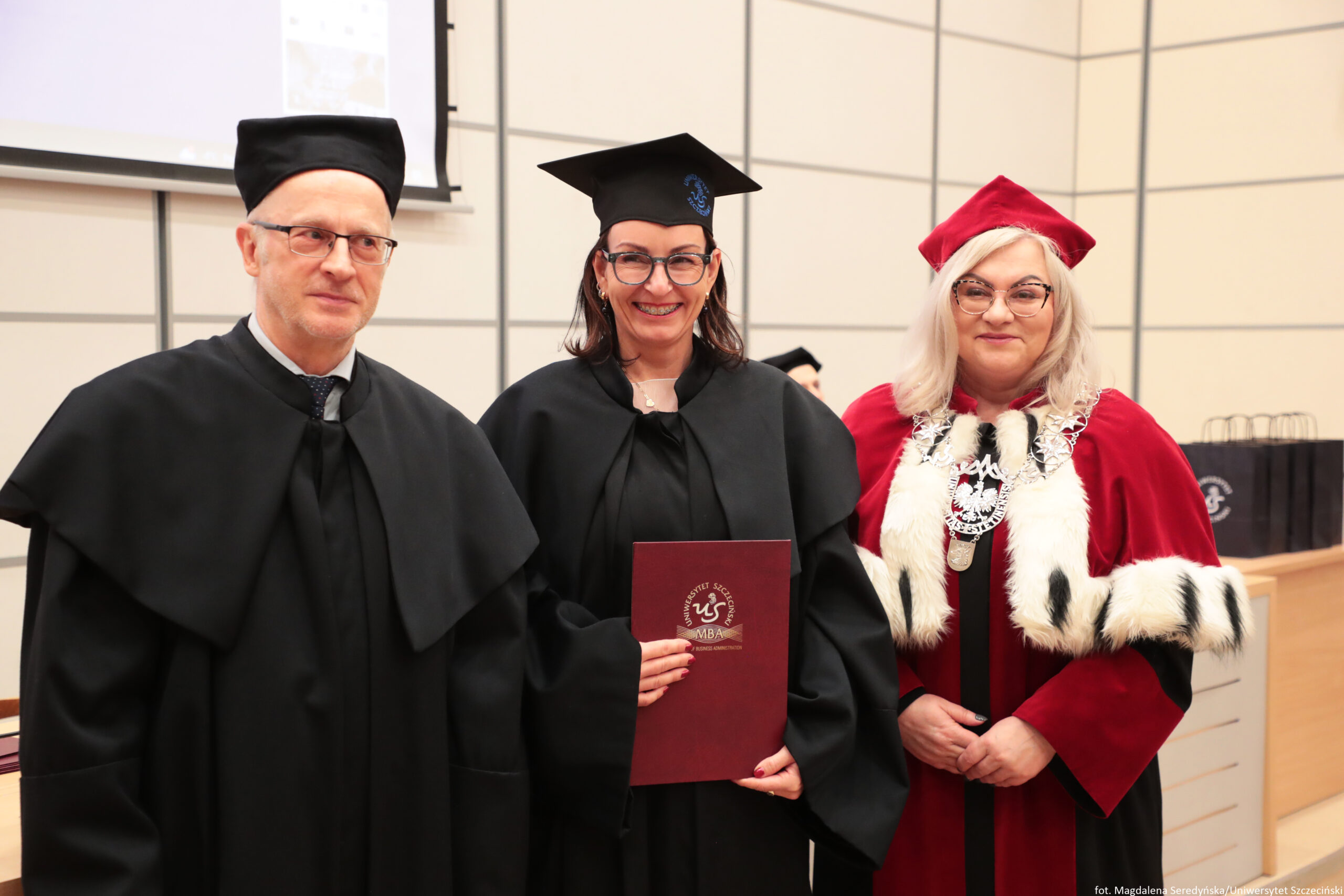 Trzy osoby w strojach akademickich podczas uroczystości, ustawione do zdjęcia w sali wykładowej. Po lewej stoi mężczyzna w czarnym todze i czarnym birecie, w okularach, z poważnym wyrazem twarzy. Na środku stoi kobieta w czarnej todze i czarnym birecie, w dużych okularach, uśmiechnięta; trzyma bordowy dyplom lub teczkę. Na okładce widać napis „UNIWERSYTET SZCZECIŃSKI” oraz „MBA” i okrągłe godło z białym symbolem. Po prawej stoi kobieta w czerwonym stroju ceremonialnym z jasnym futrzanym wykończeniem i czerwonym biretem, w okularach; na szyi ma duży łańcuch z medalionem. W tle widać jasne ściany z podziałami, fragment ekranu/projektora oraz biurko. W prawym dolnym rogu jest mały podpis: „fot. Magdalena Seredyńska/Uniwersytet Szczeciński”.