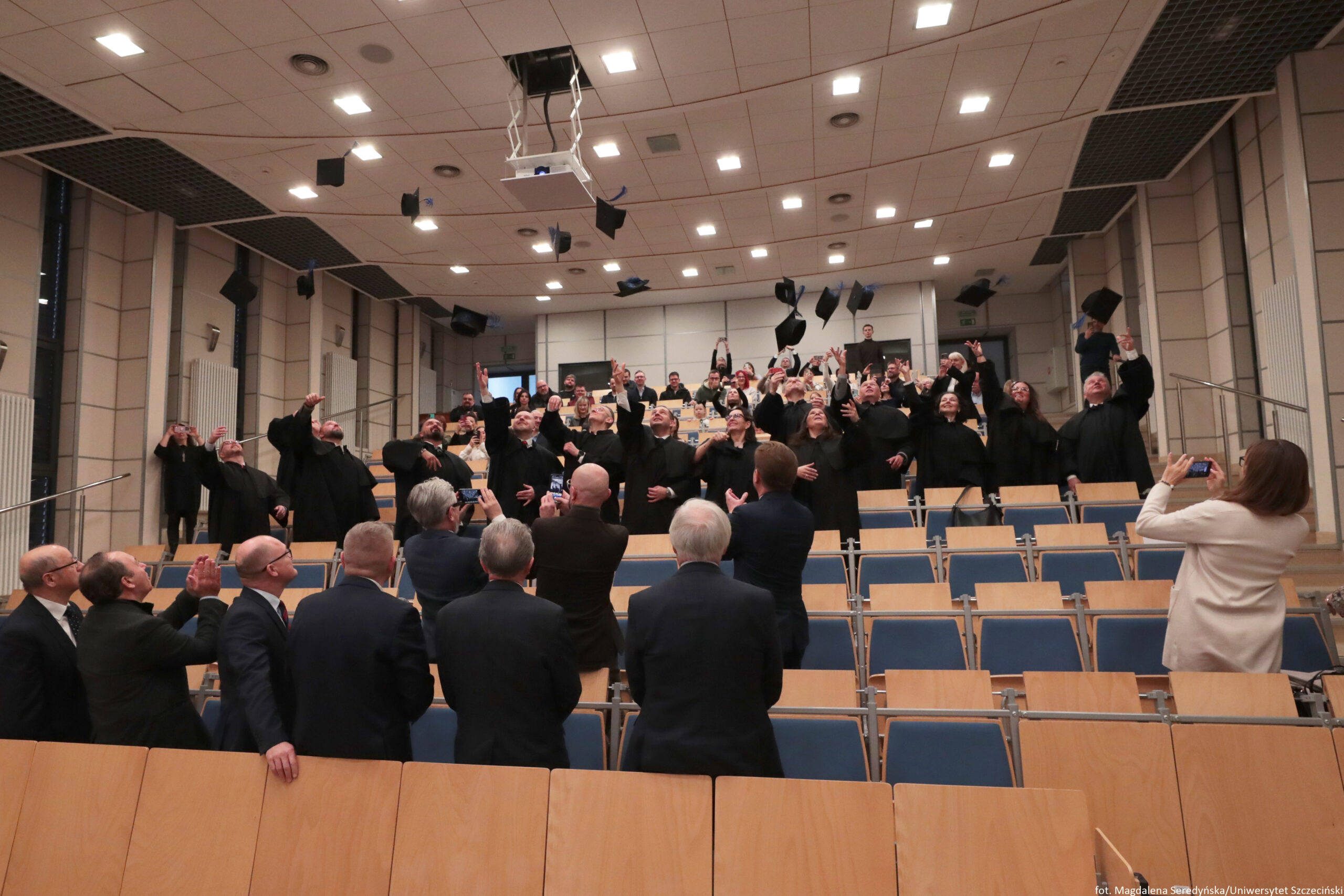 Aula wykładowa z rzędami niebieskich krzeseł i drewnianych pulpitów, pełna ludzi świętujących i podrzucających w górę czarne birety. W środkowej części sali stoi duża grupa osób w czarnych togach; wiele z nich ma uniesione ręce i patrzy w górę na lecące birety. Na pierwszym planie widać kilka osób w garniturach, część robi zdjęcia telefonami. Po prawej stronie stoi osoba w jasnym płaszczu, również fotografująca. Na suficie są liczne kwadratowe lampy, a centralnie zwisa podwieszony projektor. W prawym dolnym rogu znajduje się mały napis: „fot. Magdalena Seredyńska/Uniwersytet Szczeciński”.