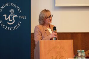 Po lewej granatowy rollup z białym okrągłym logiem Uniwersytetu Szczecińskiego. Na środku drewniana mównica, za którą stoi z mikrofonem dr hab. Adrianna Seniów, prof. US podczas III Międzynarodowego Kongresu Języka i Kultury Polskiej.