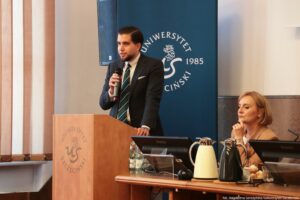 III Międzynarodowy Kongres Języka i Kultury Polskiej. Po lewej dr Michał Baran za mównicą z mikrofonem. Za nim granatowy rollup z białym okrągłym logiem Uniwersytetu Szczecińskiego. Po prawej siedzi dr hab. Adrianna Seniów, prof. US.