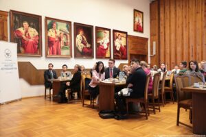 Ujęcie bokiem. Siedzący przy stołach (3 rzędy) uczestnicy III Międzynarodowego Kongresu Języka i Kultury Polskiej. W tle na ścianie portrety rektorów w gronostajach.