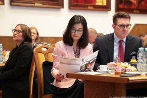 Ujęcie bokiem. Siedzący przy stołach uczestnicy III Międzynarodowego Kongresu Języka i Kultury Polskiej. Zbliżenie na 3 osoby. Kobieta w środku czyta program wydarzenia.