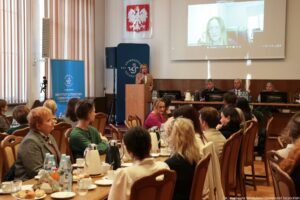 Ujęcie bokiem. Na pierwszym planie siedzący przy stołach tyłem i bokiem do kadru uczestnicy III Międzynarodowego Kongresu Języka i Kultury Polskiej. W tle po lewej granatowe rollupy – z białym okrągłym logiem Uniwersytetu Szczecińskiego i logiem Instytutu Literatury i Nowych Mediów, mównica, za którą stoi z mikrofonem gospodyni spotkania, nad nią godło Polski, dalej stół prezydialny, za którym siedzą rektorzy. U góry na ścianie ekran z prelegentką.