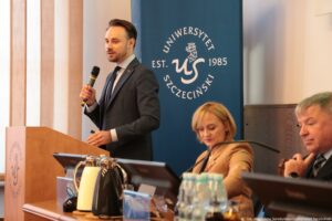 III Międzynarodowy Kongres Języka i Kultury Polskiej. Ujęcie bokiem. Od lewej: Dr Damian Syjczak (za mównicą z mikrofonem), dr hab. Adrianna Seniów, prof. US i prof. Andrzej Skrendo. W tle granatowy rollup z białym okrągłym logiem Uniwersytetu Szczecińskiego.