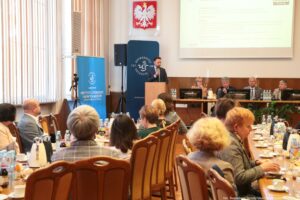 Na pierwszym planie siedzący przy stołach tyłem i bokiem do kadru uczestnicy III Międzynarodowego Kongresu Języka i Kultury Polskiej. W tle po lewej granatowe rollupy – z białym okrągłym logiem Uniwersytetu Szczecińskiego i logiem Instytutu Literatury i Nowych Mediów, mównica, za którą stoi z mikrofonem dr Damian Syjczak, nad nim godło Polski, dalej stół prezydialny, za którym siedzą rektorzy. U góry na ścianie ekran.