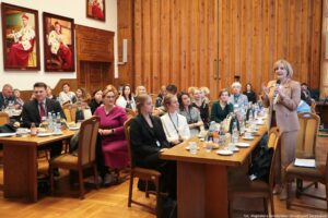Ujęcie bokiem. Siedzący przy stołach (3 rzędy) uczestnicy III Międzynarodowego Kongresu Języka i Kultury Polskiej. Po prawej stoi mikrofonem gospodyni spotkania. Po lewej w tle na ścianie portrety rektorów w gronostajach.